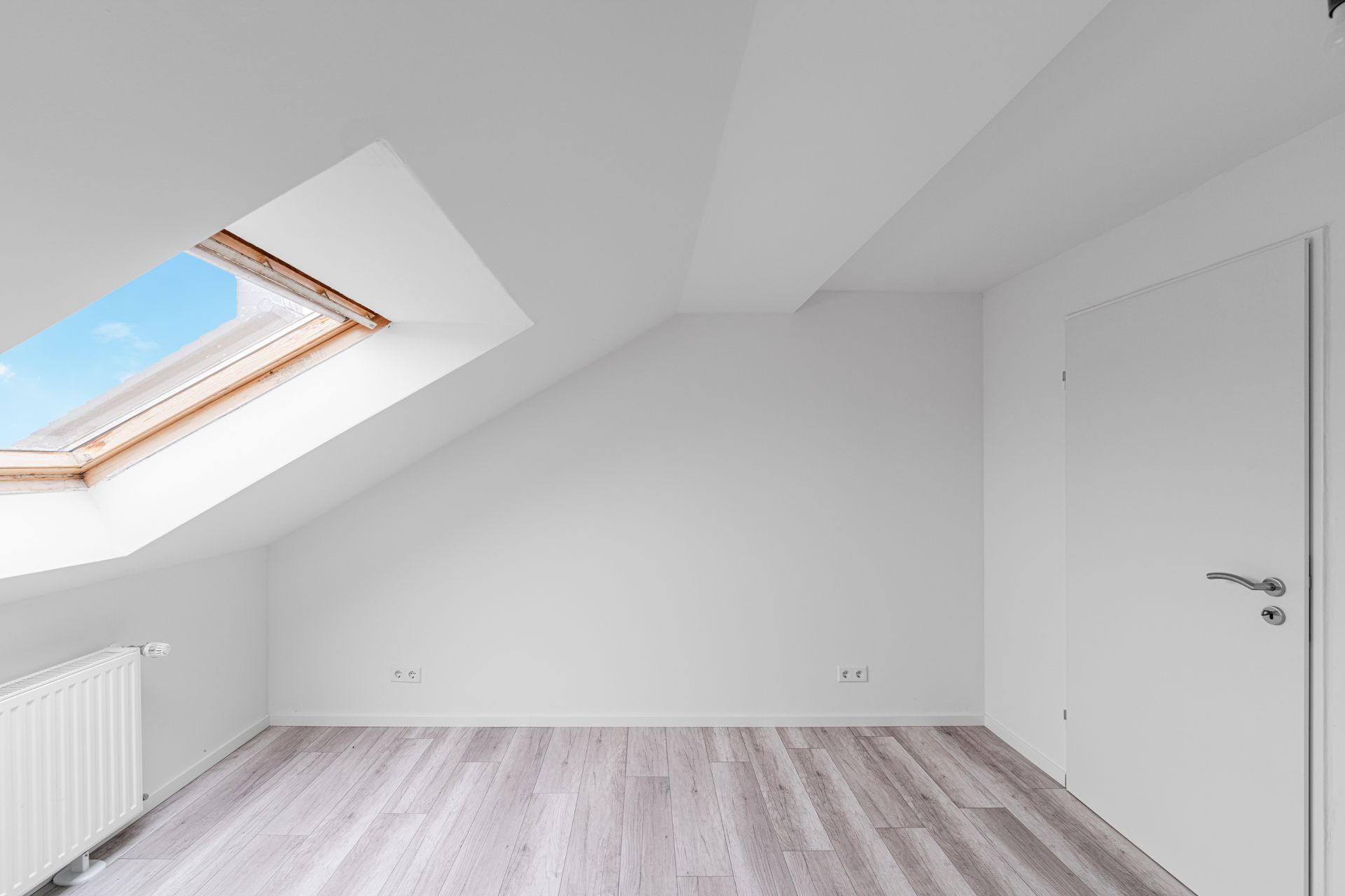 Helles Zimmer mit Dachschräge, großem Dachfenster, weißer Tür und hellgrauem Holzboden.