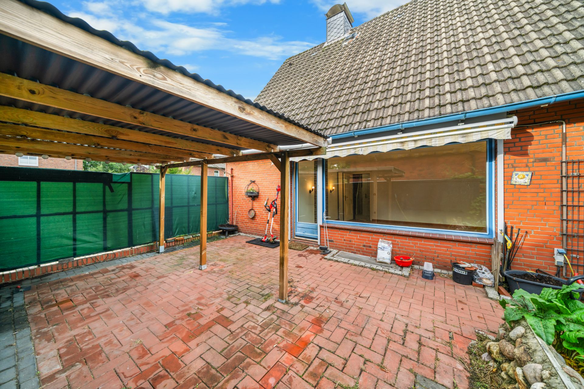 Gepflasterter Innenhof mit Überdachung, Sichtschutzzaun und Gartenwerkzeugen neben einem Backsteinhaus.