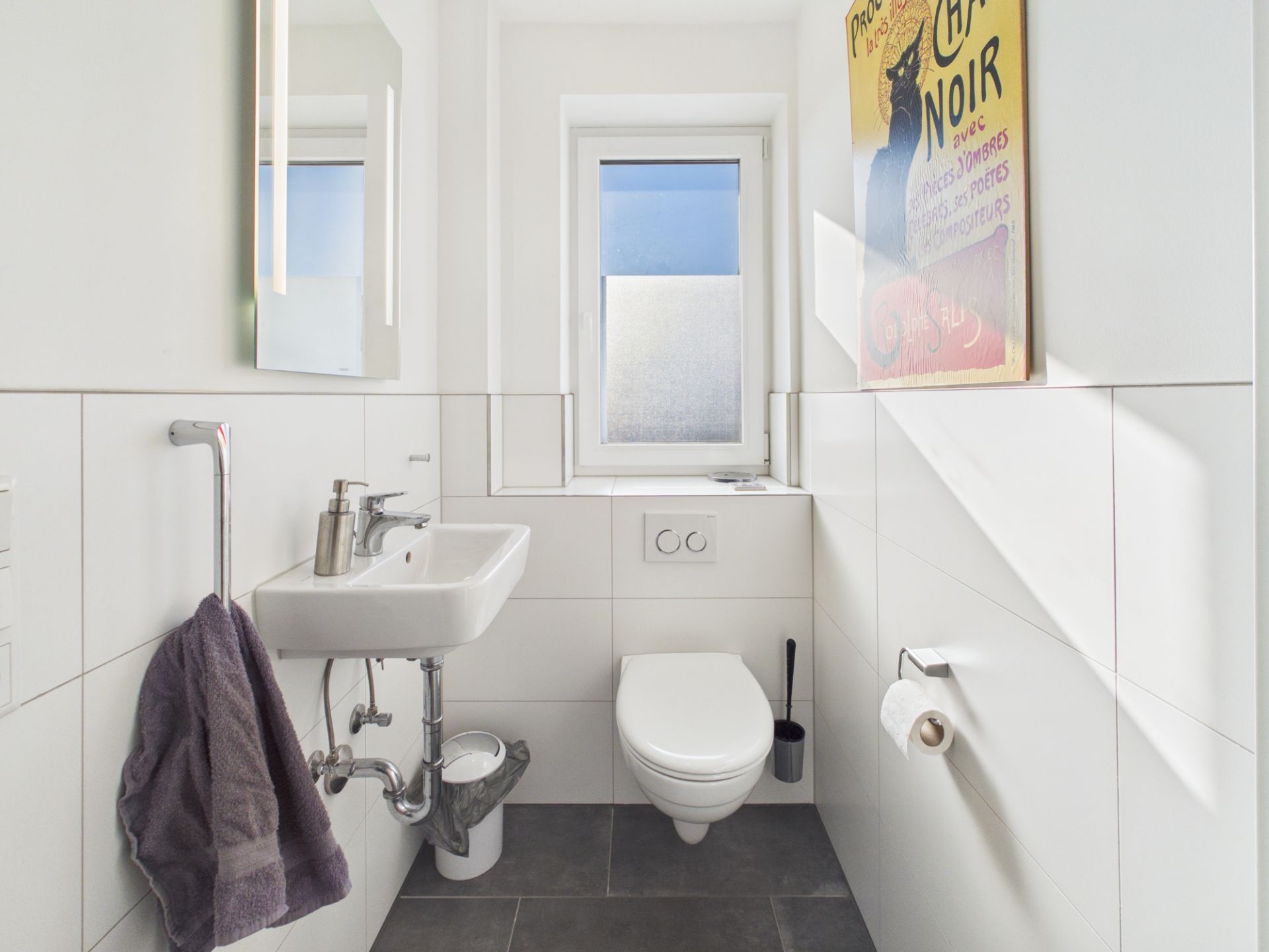 Kleines, weiß gefliestes Bad mit Wandtoilette, Waschbecken, Fenster und einem Handtuch an der Seite.