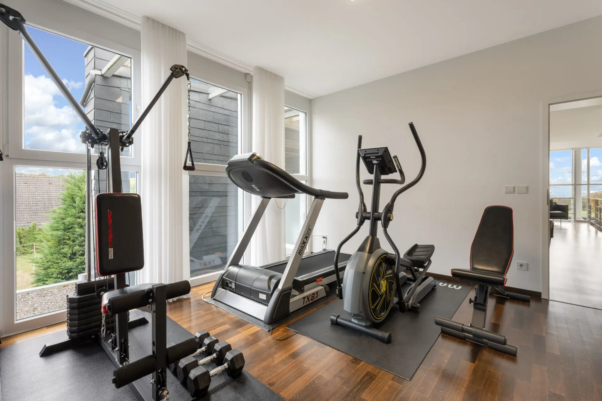 Helles Fitnesszimmer mit Kraftstation, Laufband, Crosstrainer und Hanteln auf Holzboden.