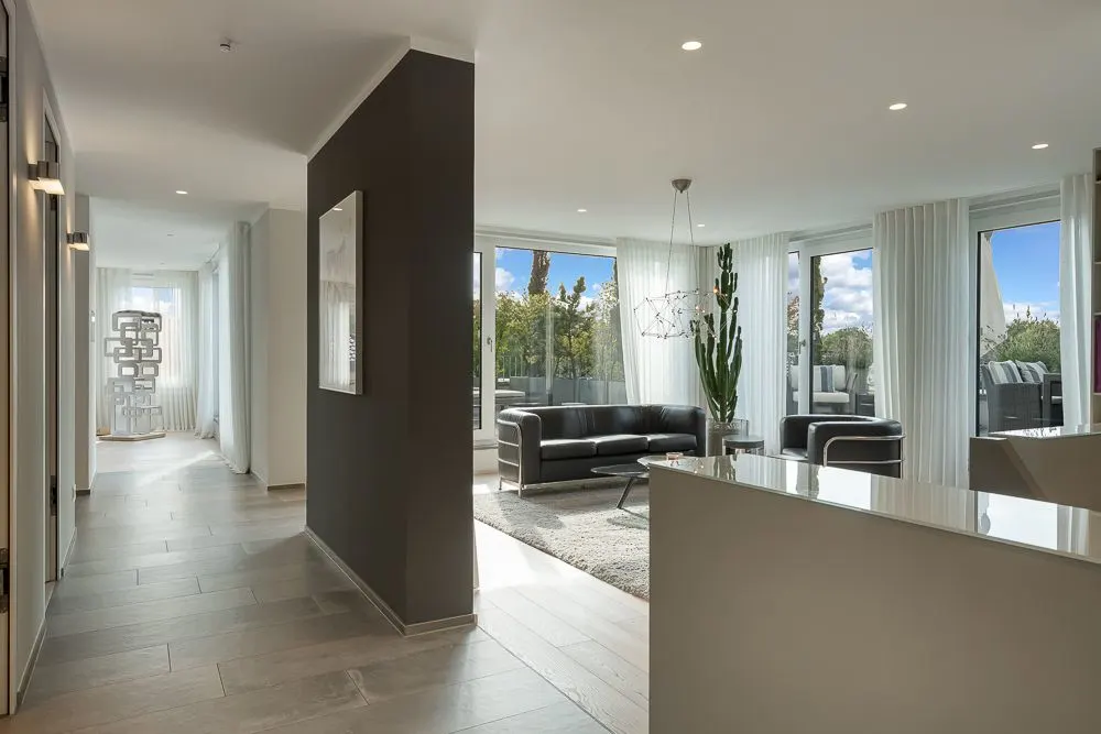 Modernes Wohnzimmer mit großen Fenstern, schwarzem Sofa und Pflanzen, Flur im Vordergrund.
