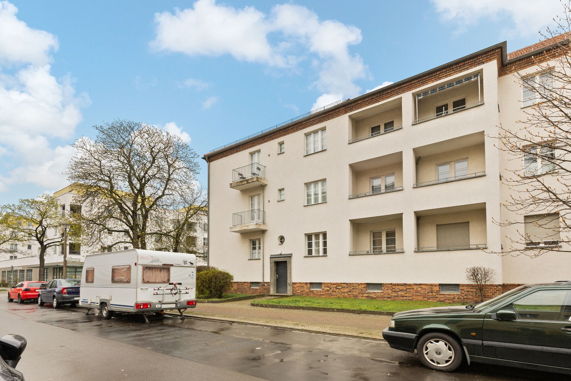 Mehrstöckiges Wohnhaus mit Balkonen, geparkte Autos und Wohnwagen am Straßenrand.