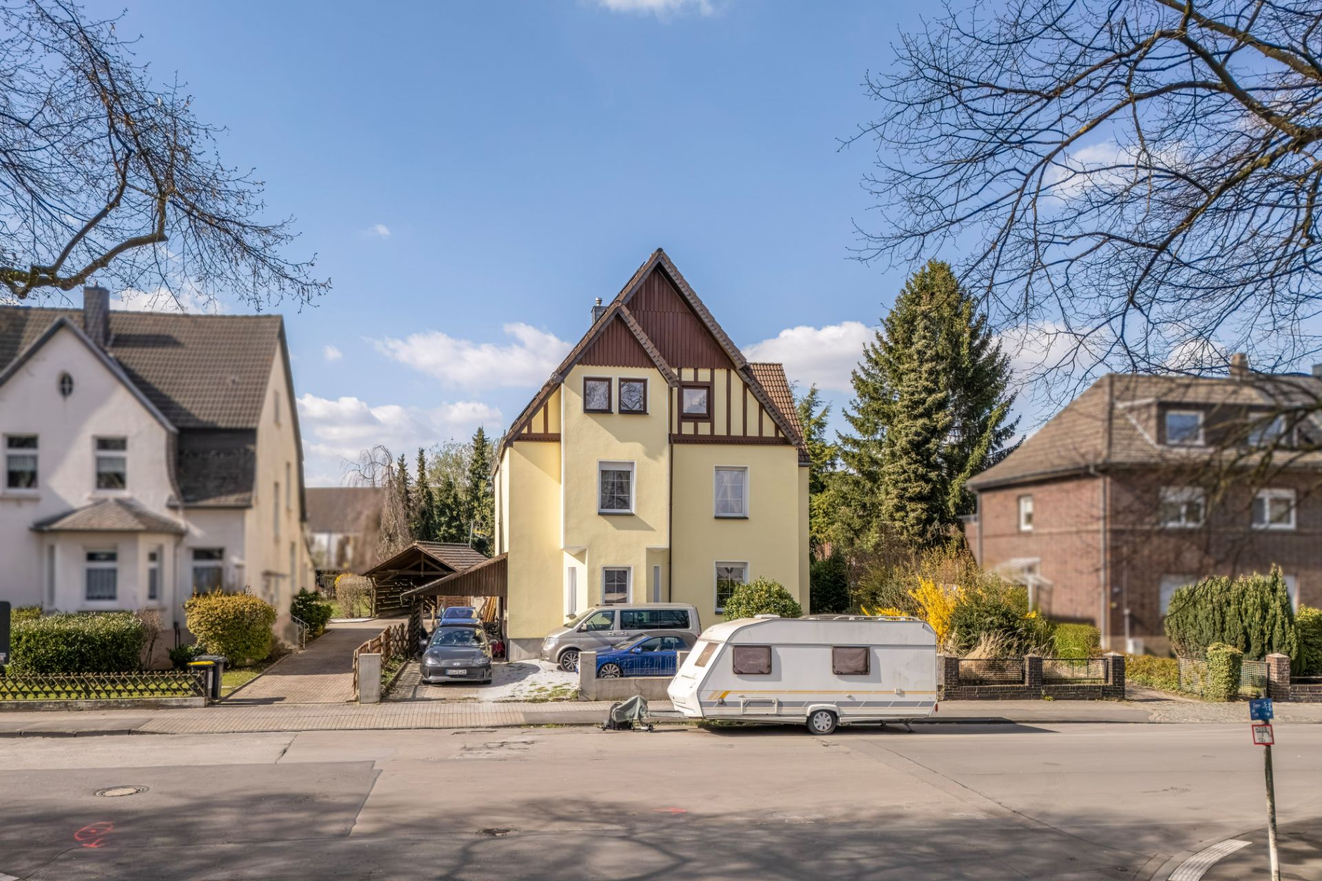 Gelbes Wohnhaus mit spitzem Dach, mehreren Autos und einem Wohnwagen davor auf der Straße.