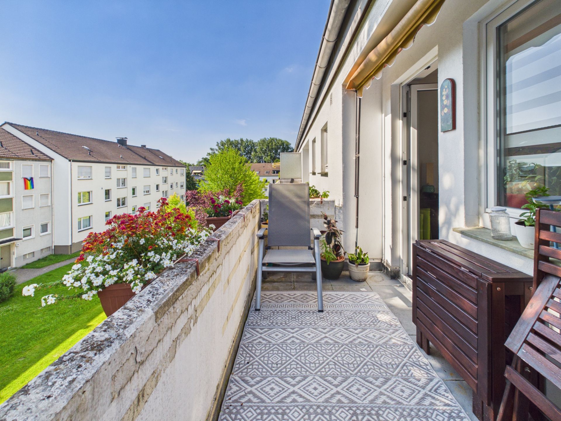 Balkon mit Blumen, Stuhl, Holztisch und Blick auf einen grünen Innenhof bei sonnigem Wetter.