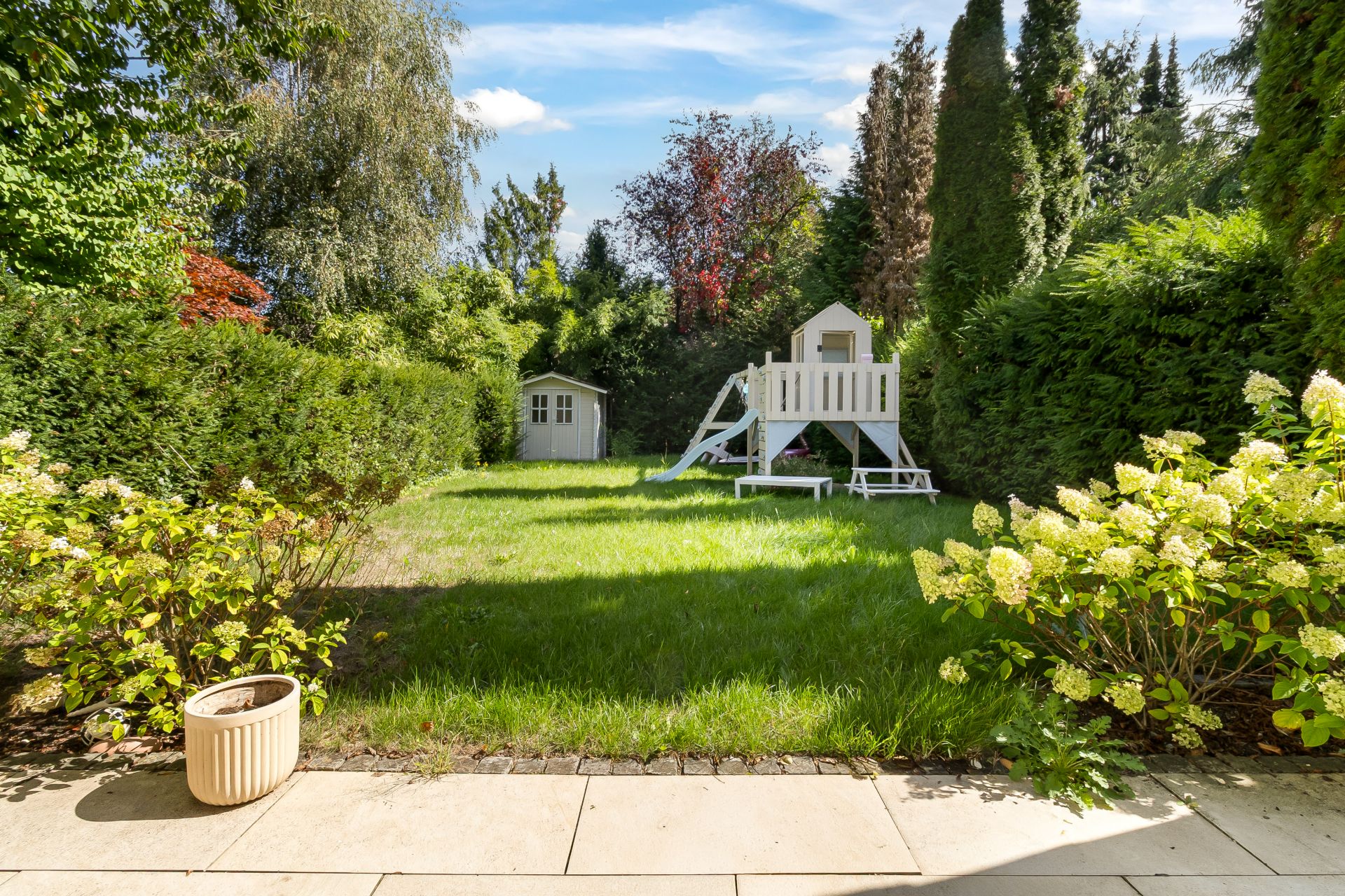 Garten mit Spielturm, Rutsche, Picknicktisch und kleinem Gartenhaus, umgeben von Büschen und Bäumen.