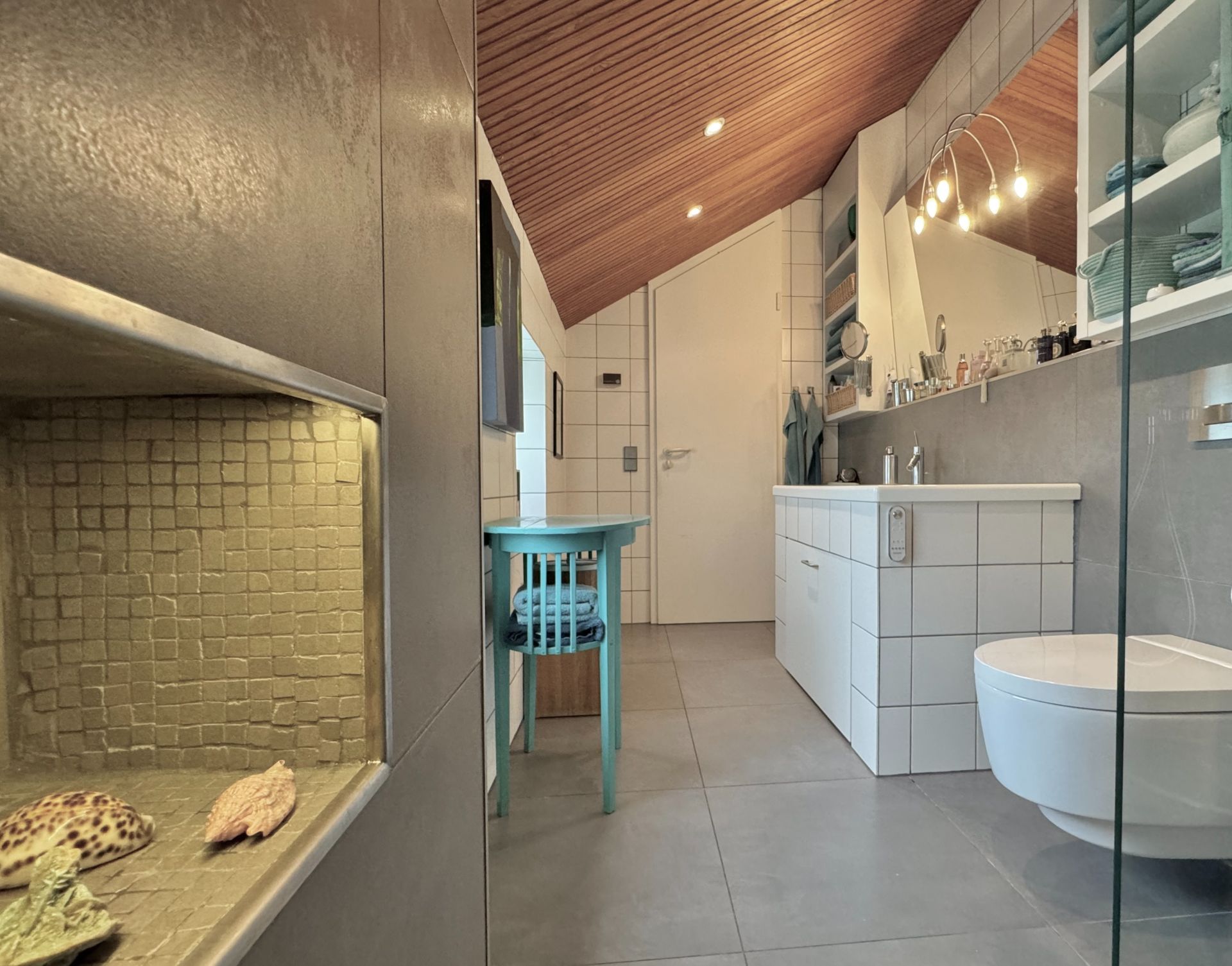Modernes Badezimmer mit Holzdecke, weißem Waschbecken, Wandregalen und rundem Tisch mit Stuhl.