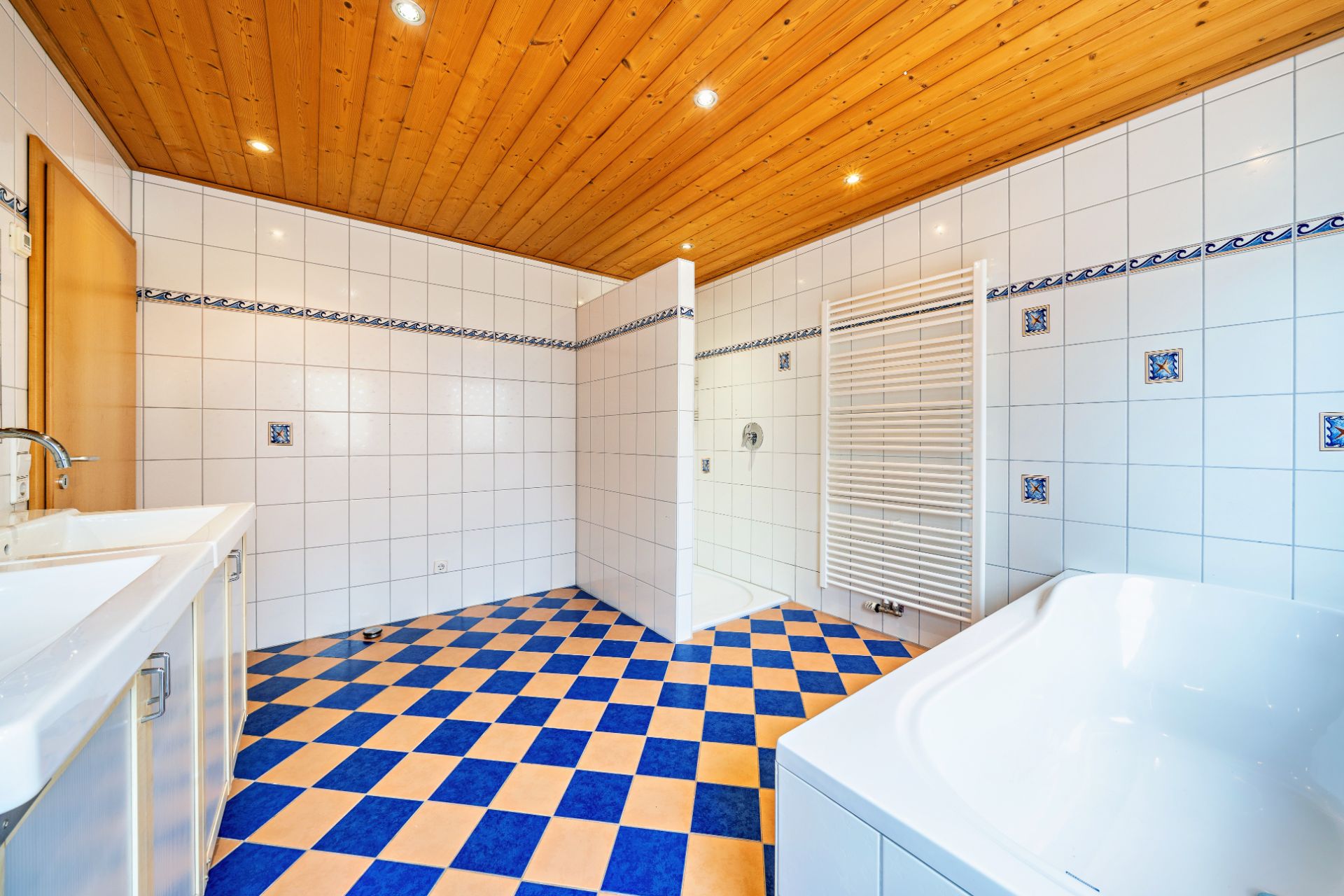 Helles Bad mit blau-gelbem Fliesenboden, Badewanne, Doppelwaschbecken und Holzdecke.