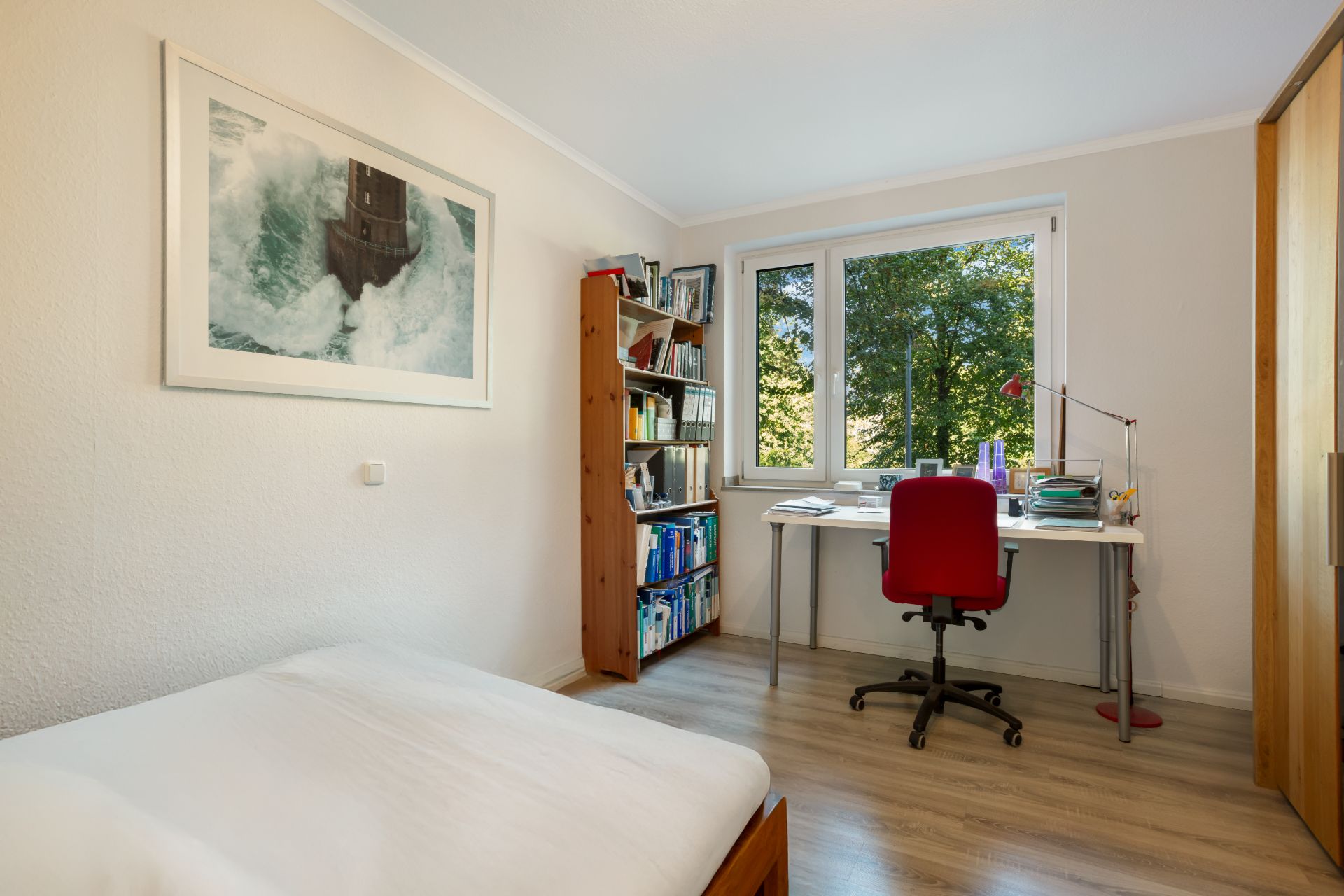 Helles Schlafzimmer mit Schreibtisch, rotem Stuhl, Regal und Fenster mit Blick ins Grüne.
