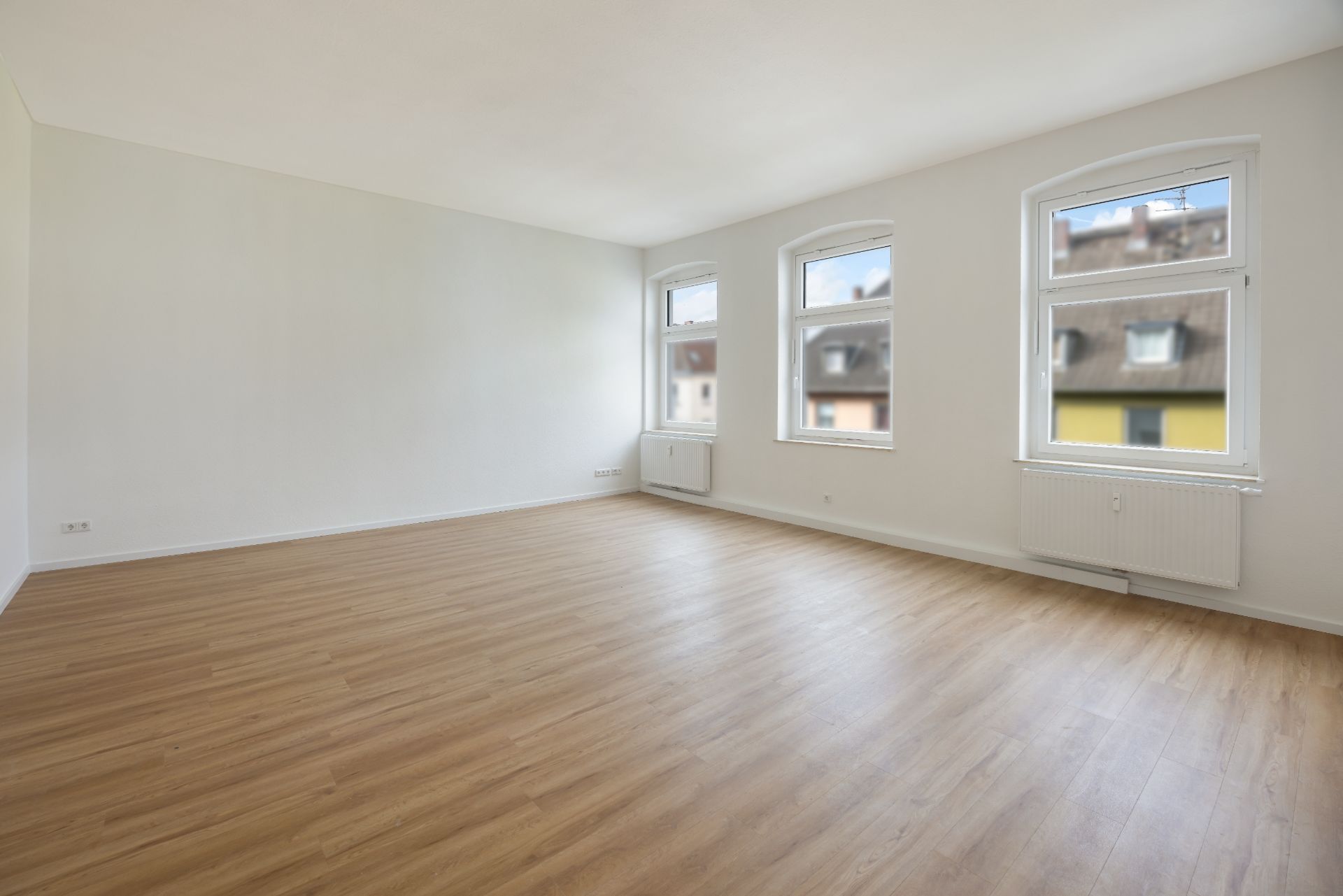 Leerer Raum mit Holzfußboden und drei großen Fenstern an einer weißen Wand.