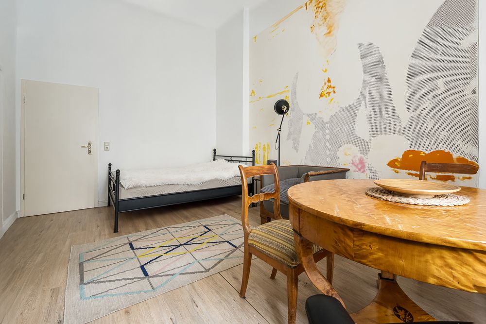 Helles Zimmer mit Holztisch, Stühlen, Einzelbett, Teppich und bunter Wandkunst.