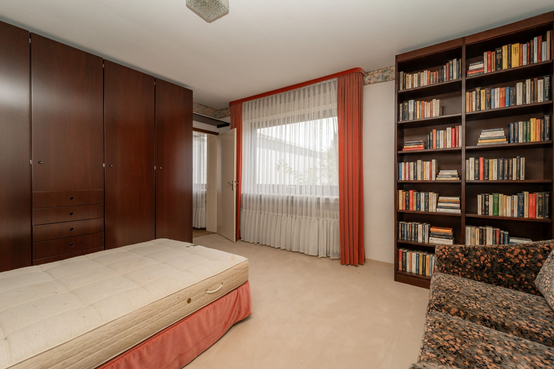 Schlafzimmer mit großem Schrank, Bett, Bücherregal und einem Fenster mit Vorhängen.
