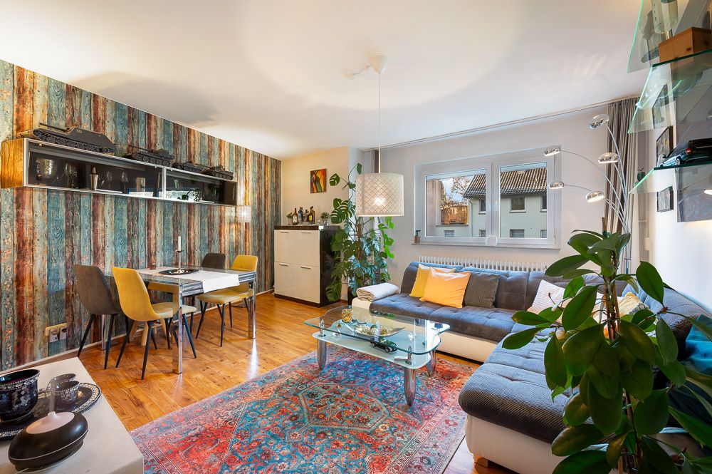 Modernes Wohnzimmer mit Ecksofa, Glastisch, buntem Teppich und Essbereich mit vier Stühlen.