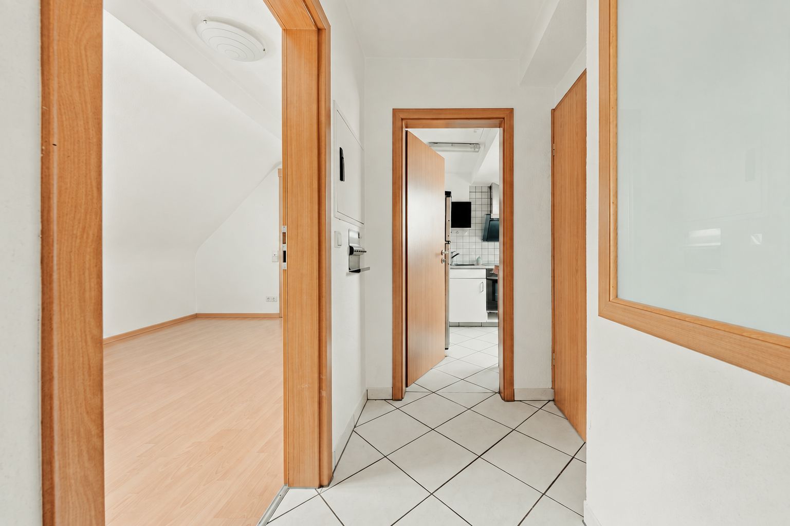Heller Flur mit Türen zu einem leeren Zimmer und einer Küche mit weißen Fliesen.
