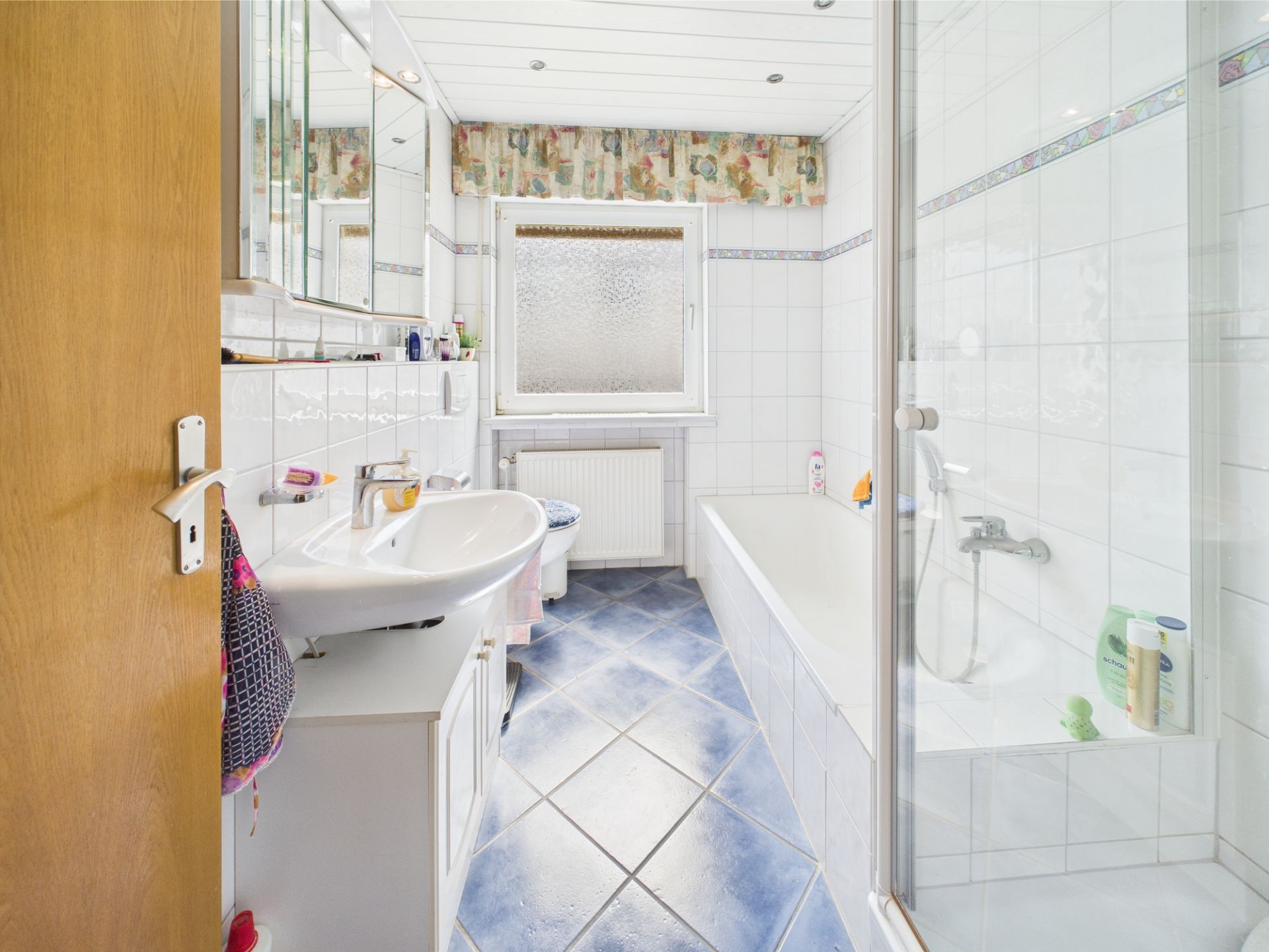 Helles Badezimmer mit Dusche, Badewanne, Waschbecken und Fenster, blauer Fliesenboden.