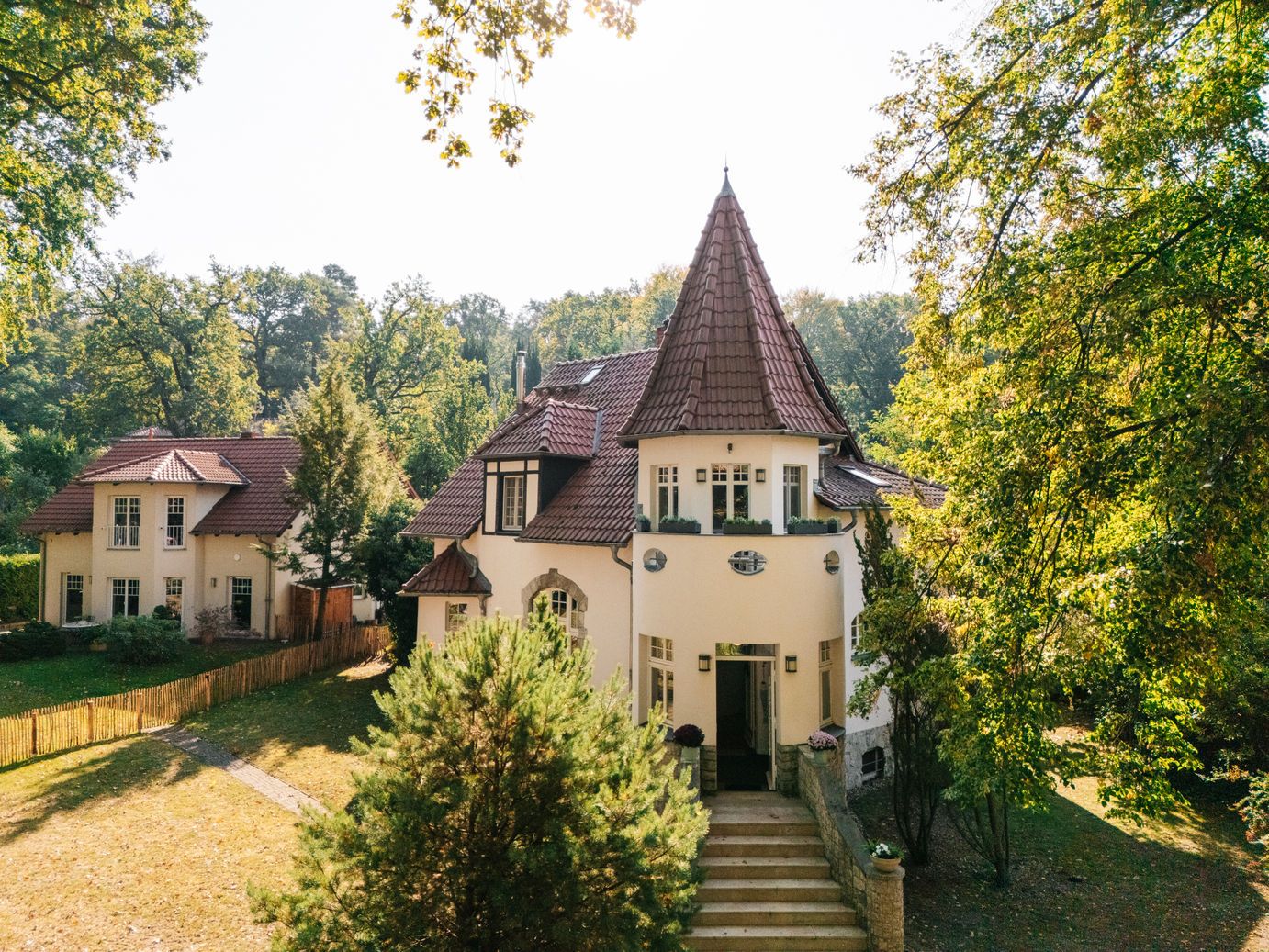 Großes Wohnhaus mit Turmdach, rotem Ziegeldach und Garten, umgeben von Bäumen.