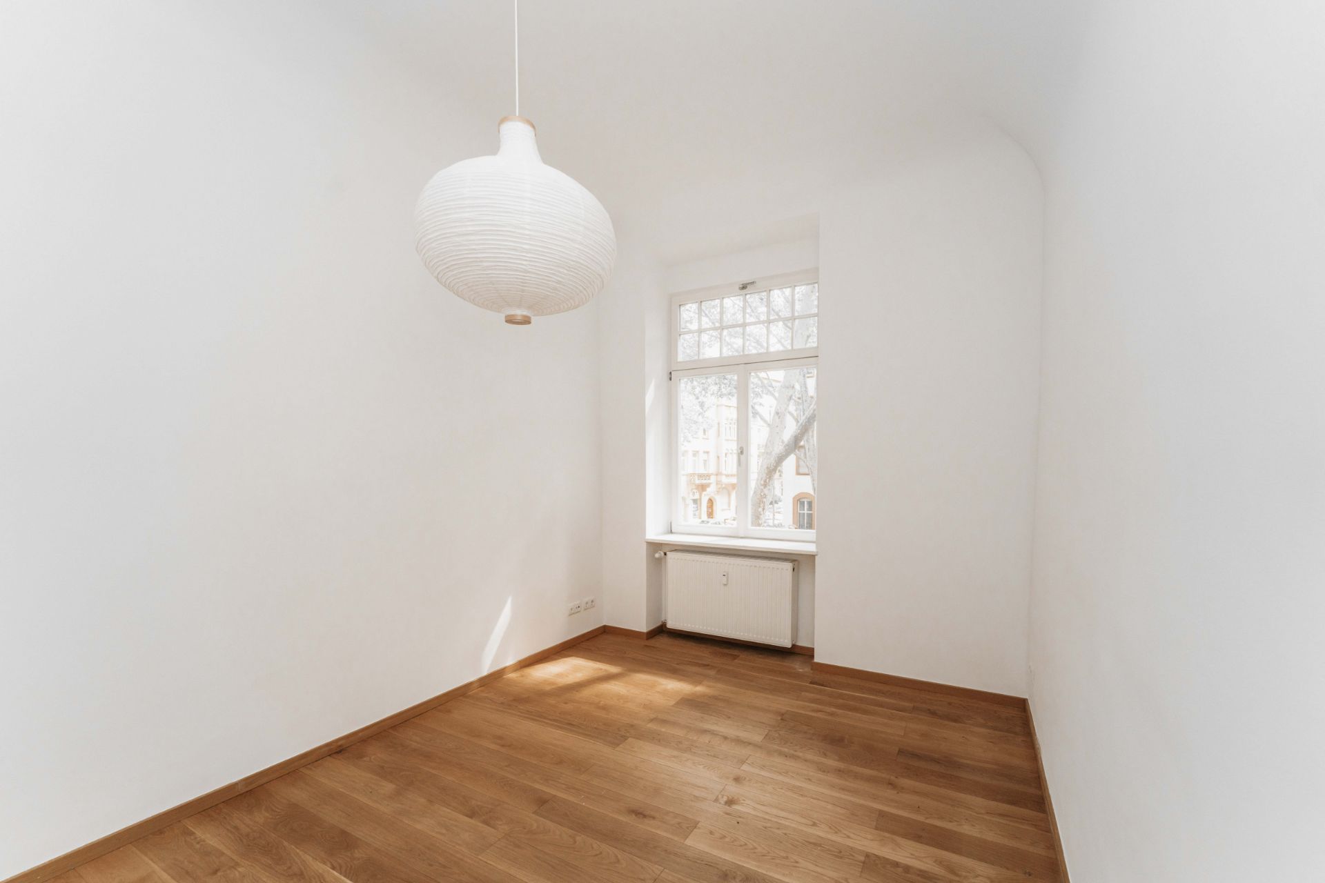 Heller leerer Raum mit Holzboden, weißer Wand, Fenster und runder Deckenlampe.