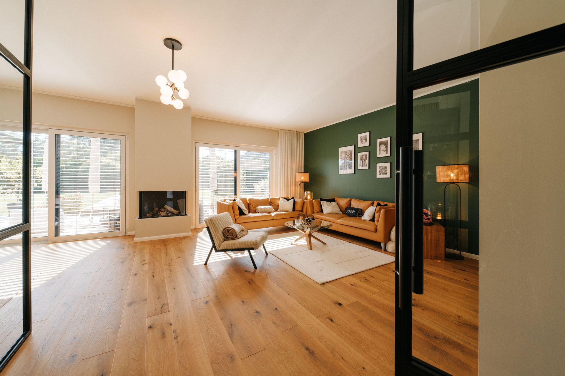 Helles Wohnzimmer mit Holzfußboden, Kamin, großer Sofa-Ecke und grüner Wand mit Bildern.