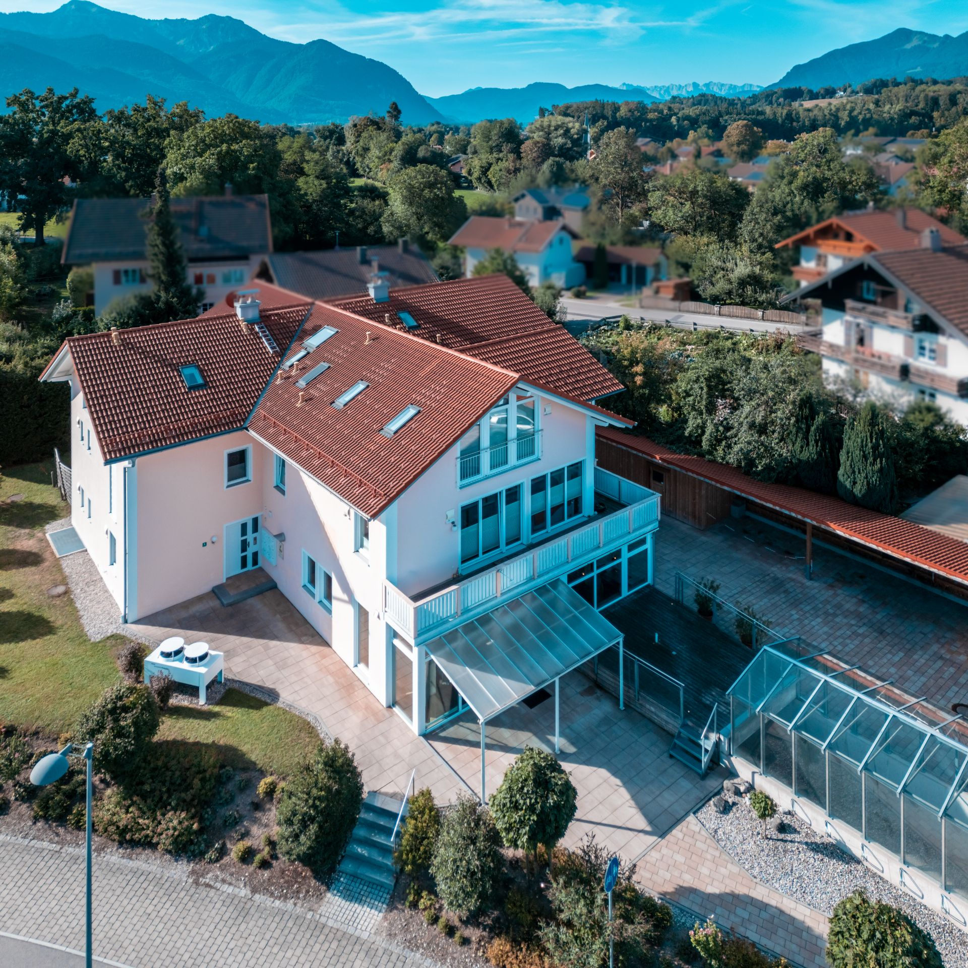 Modernes Einfamilienhaus mit rotem Dach, großem Balkon und Garten, umgeben von Bäumen und Bergen.