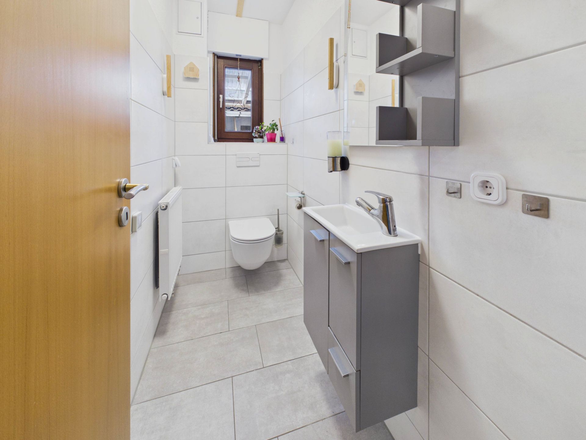 Helles Badezimmer mit Waschbecken, Spiegel, Hängeschrank, WC und Fenster.