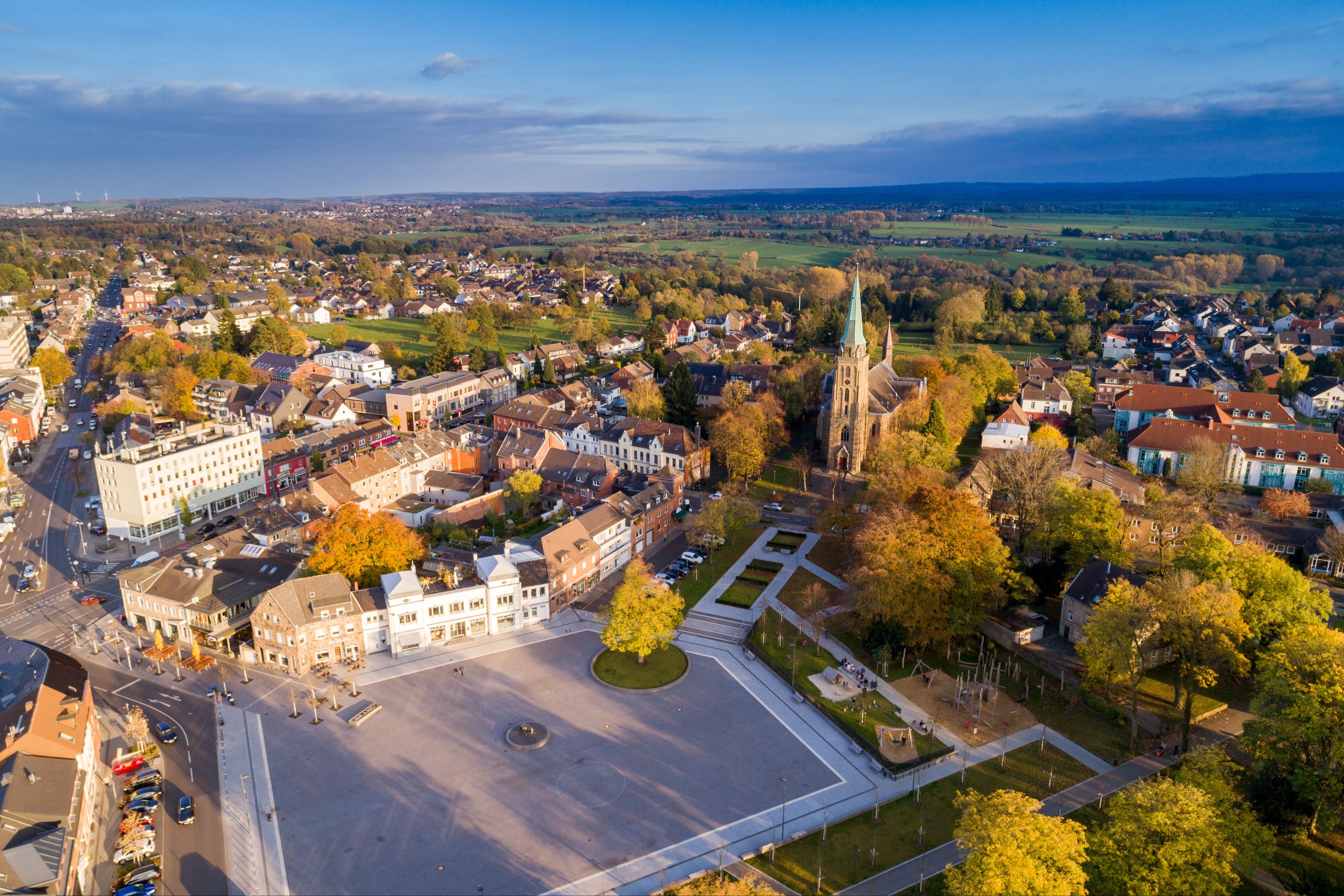 Luftaufnahme eines Stadtplatzes mit umliegenden Häusern, Bäumen und einer großen Kirche im Zentrum.