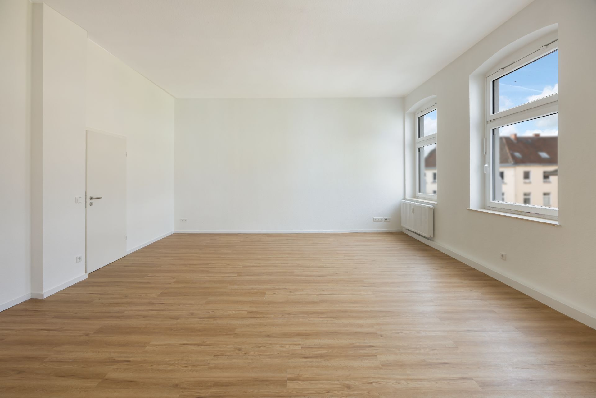 Leerer Raum mit hellem Holzboden, weißen Wänden und zwei großen Fenstern.