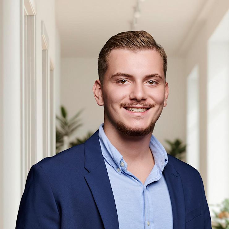 Junger Mann im blauen Anzug steht in einem hellen Flur mit Pflanzen im Hintergrund.