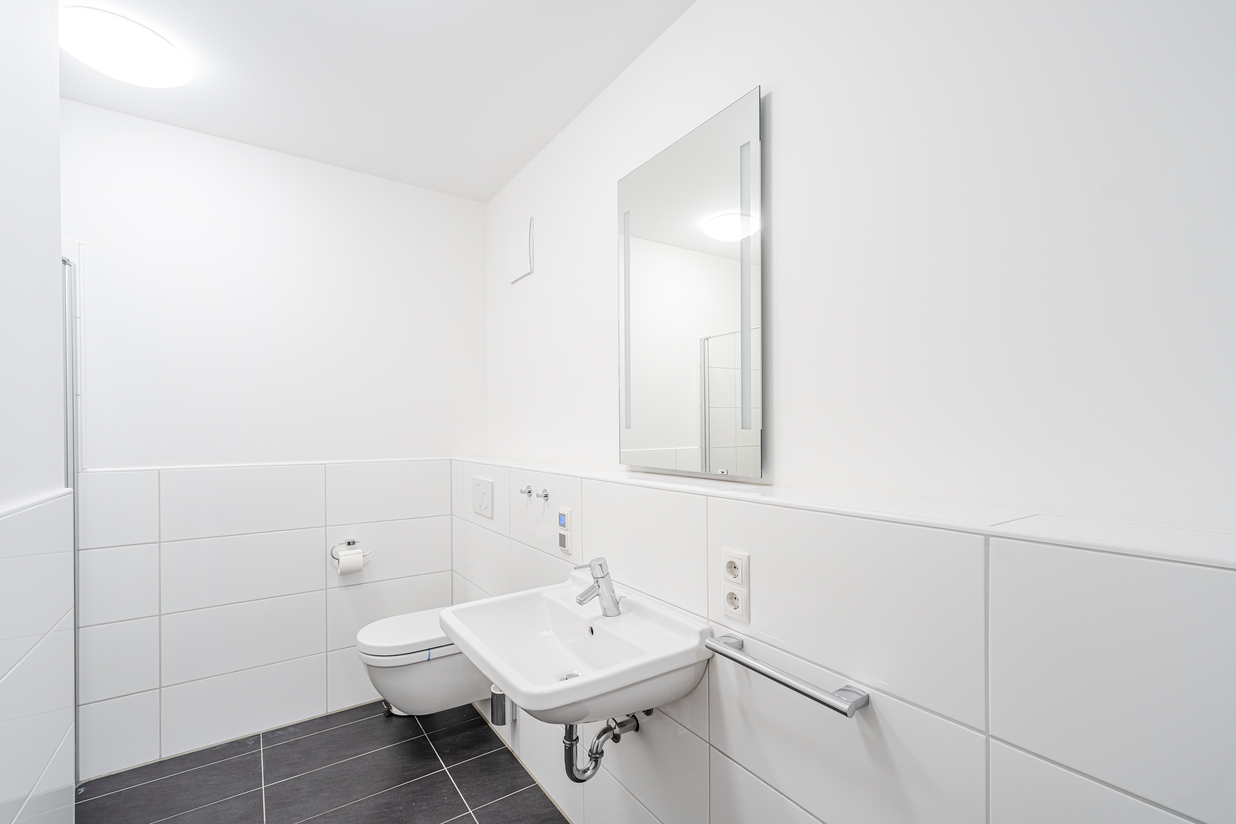Modernes, weiß gefliestes Badezimmer mit Wandtoilette, Waschbecken und Spiegel.