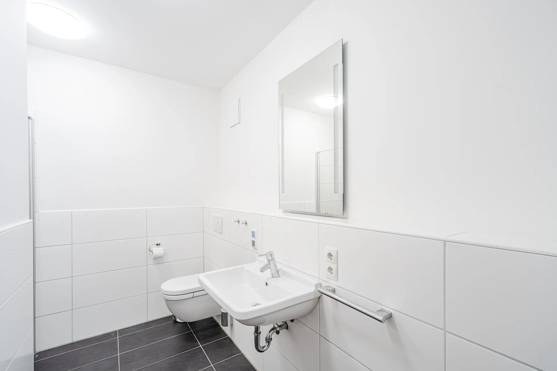 Modernes, weiß gefliestes Badezimmer mit Wandtoilette, Waschbecken und Spiegel.