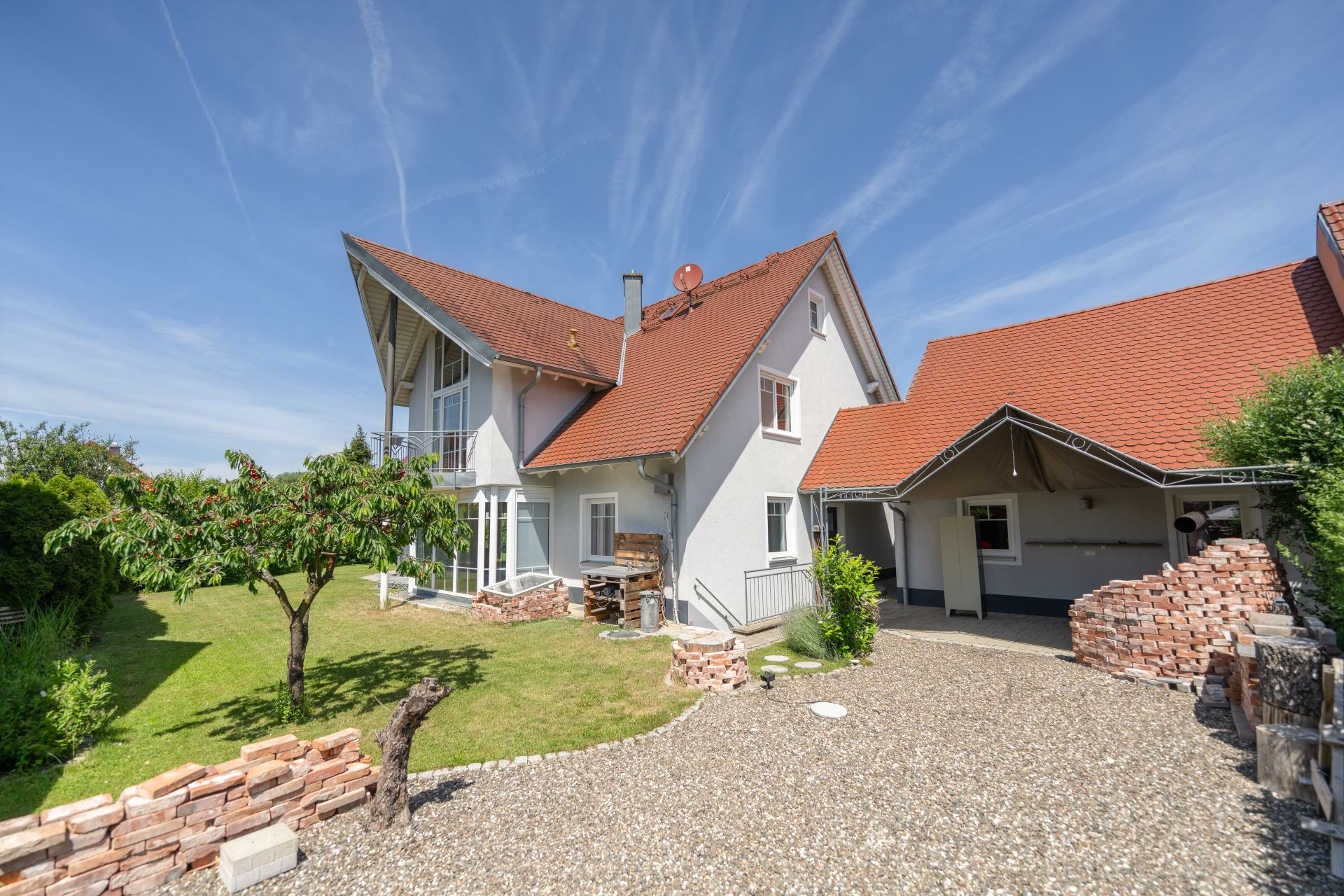 Modernes Wohnhaus mit rotem Dach, Garten, Kiesweg und gemauerten Beeten bei sonnigem Wetter.