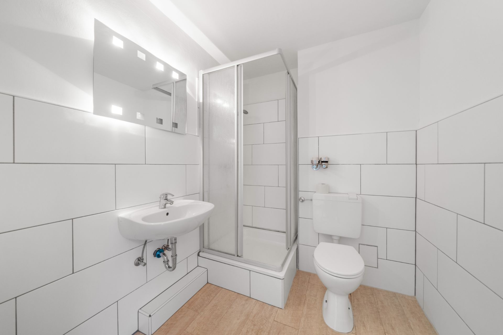 Helles Bad mit Dusche, Waschbecken, Spiegel und Toilette, weiße Fliesen an den Wänden.