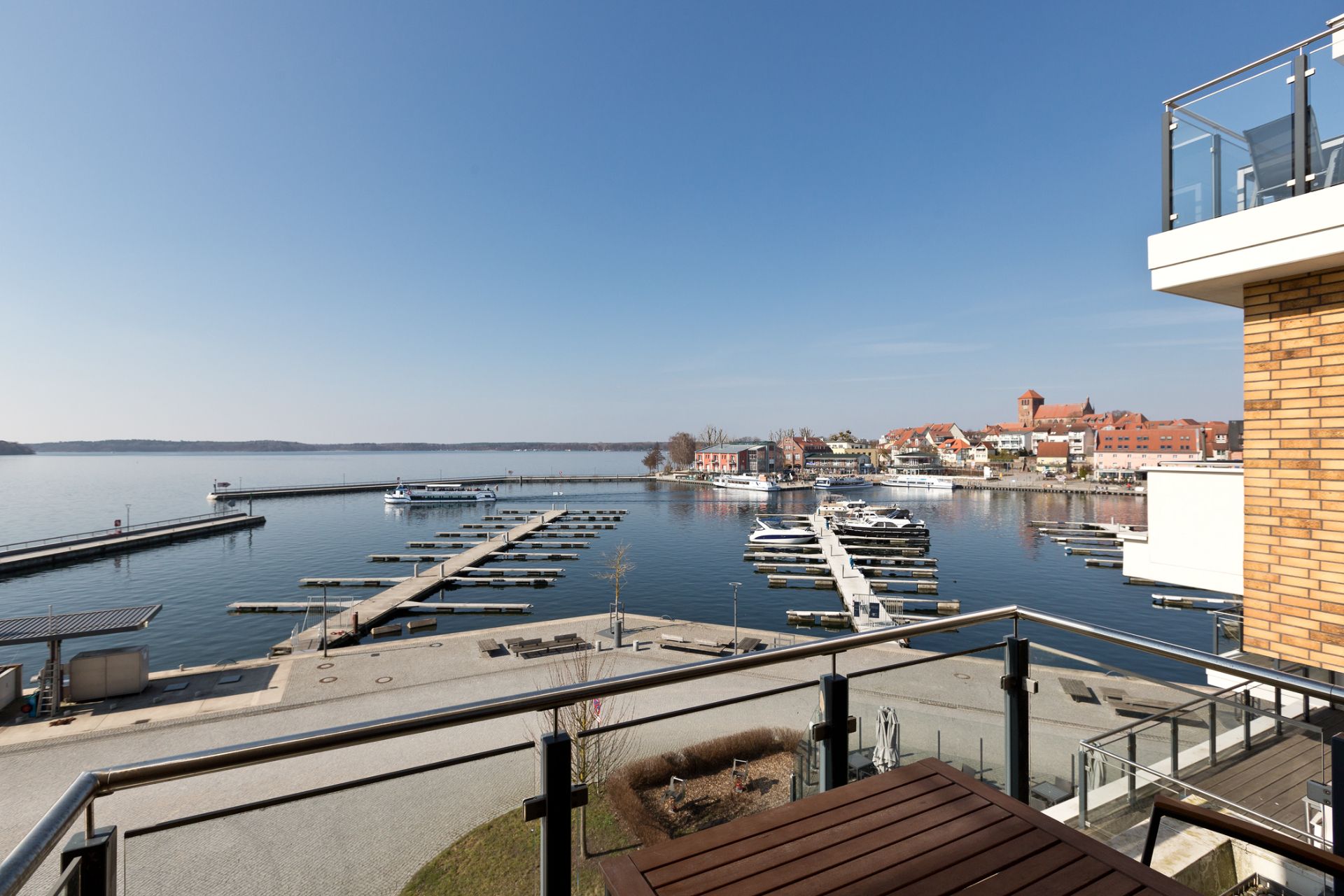 Blick von einem Balkon auf einen Yachthafen und eine Uferpromenade bei sonnigem Wetter.