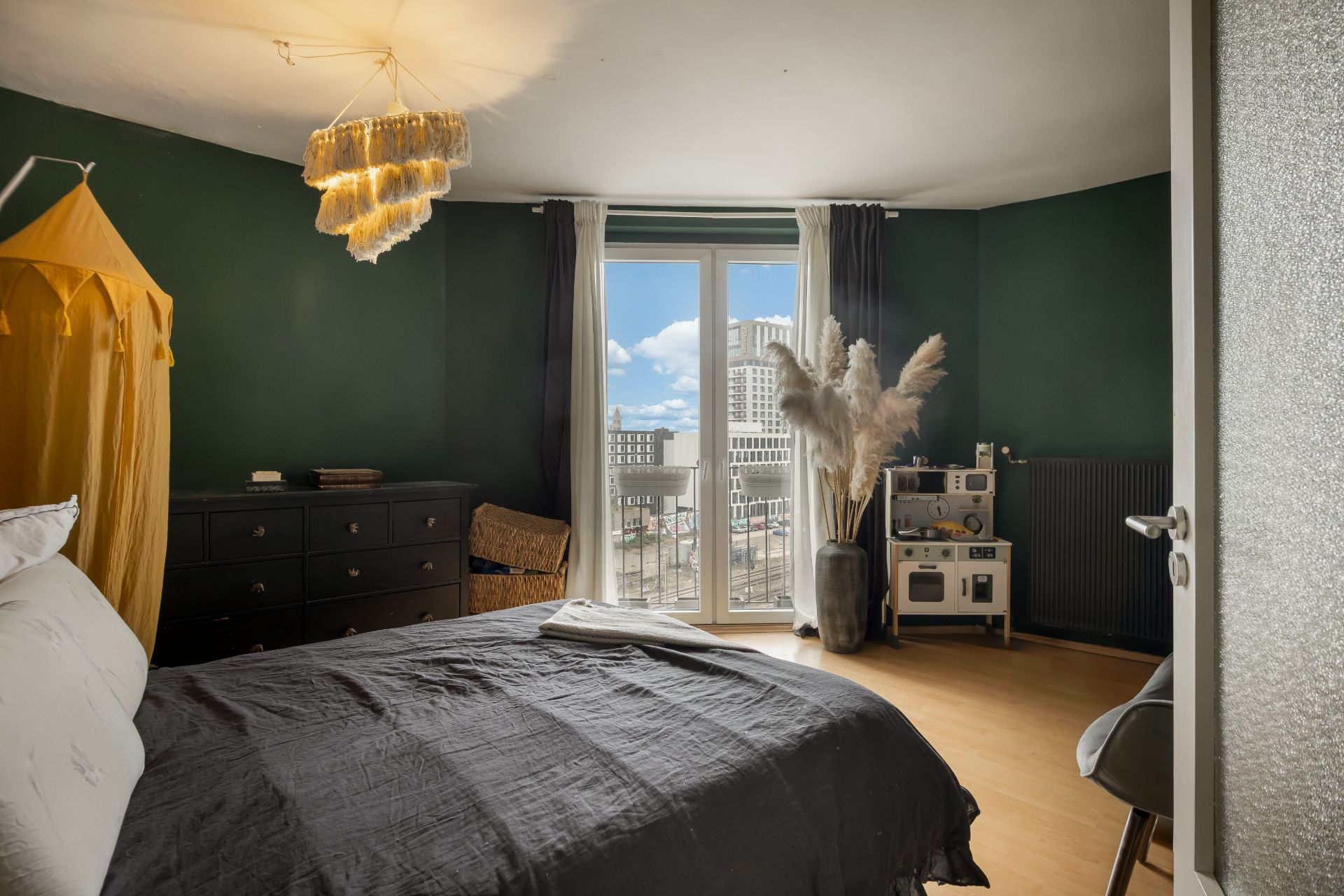 Schlafzimmer mit großem Bett, Kommode, Spielküche, und Fenster mit Blick auf Stadtgebäude.