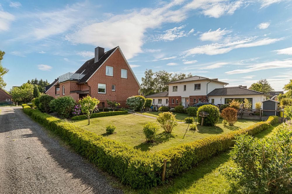 Zwei Wohnhäuser mit gepflegtem Garten und Hecke an einer Kiesstraße bei sonnigem Wetter.
