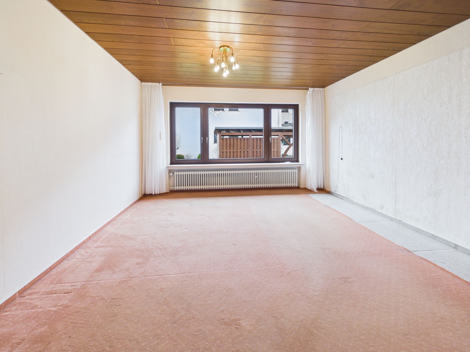 Leeres Zimmer mit großem Fenster, Vorhängen, Holzdecke und Teppichboden.