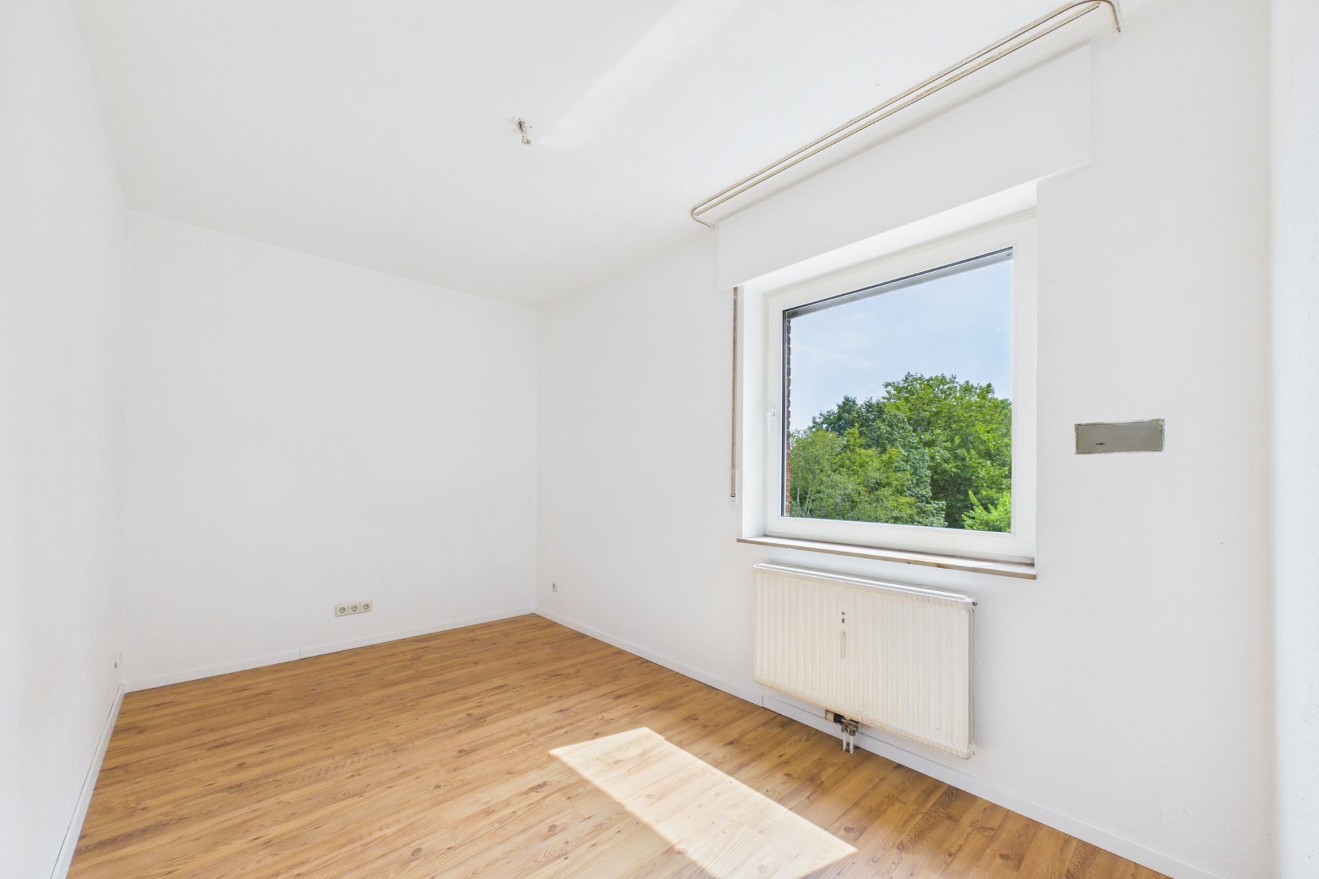 Helles, leeres Zimmer mit Holzfußboden und großem Fenster mit Blick ins Grüne.