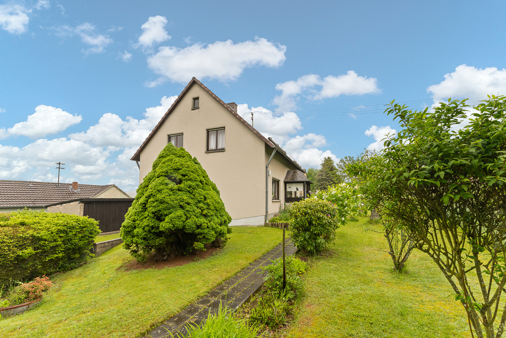 Einfamilienhaus mit Satteldach, umgeben von grünem Garten und Sträuchern bei blauem Himmel.