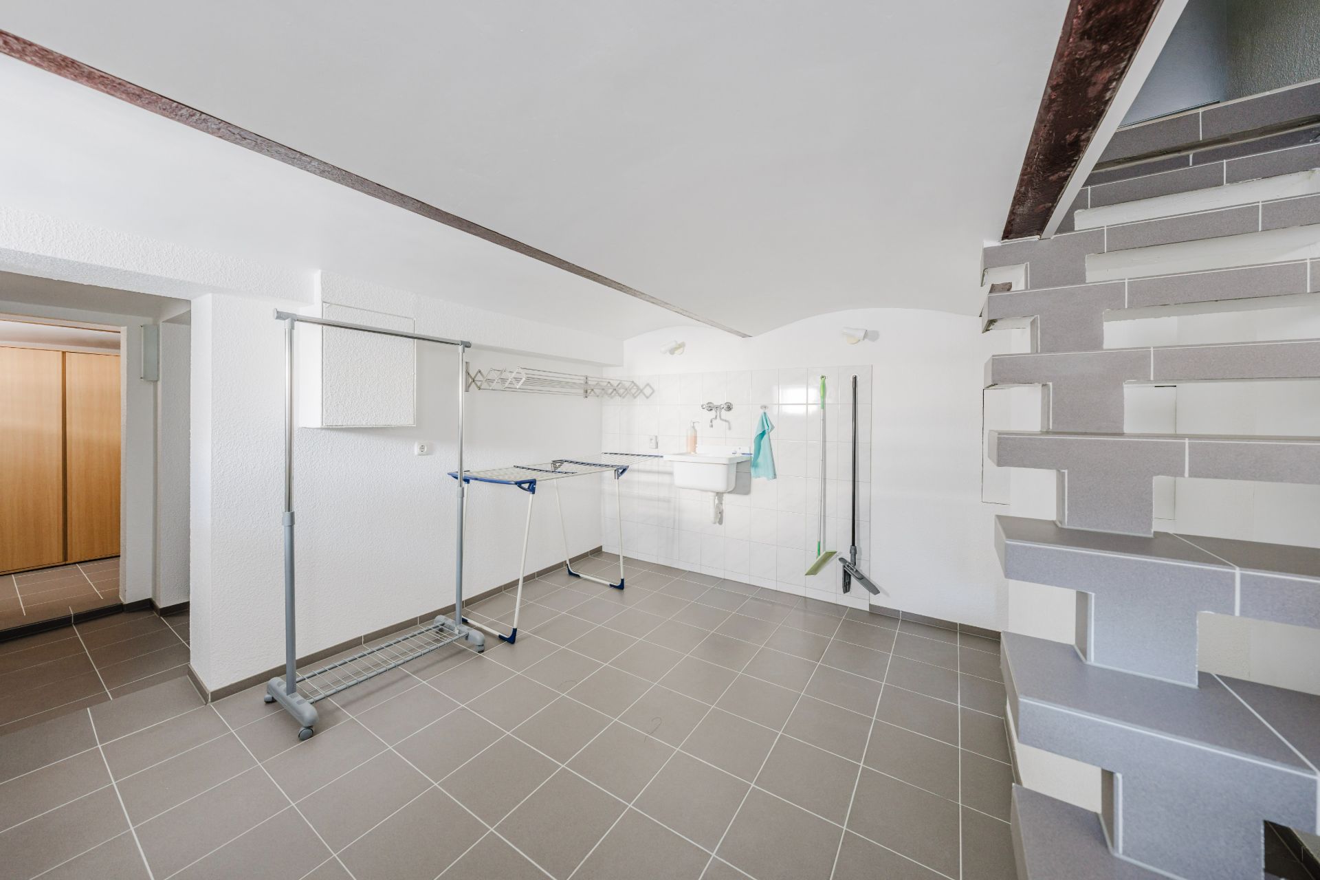Heller Kellerraum mit Fliesenboden, Wäscheständer, Waschbecken und Treppe.