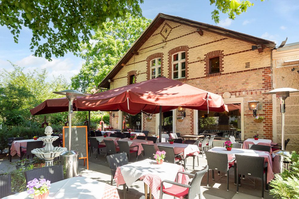 Sonniger Außenbereich mit Tischen, Stühlen, großen roten Sonnenschirmen und alter Backsteinfassade.