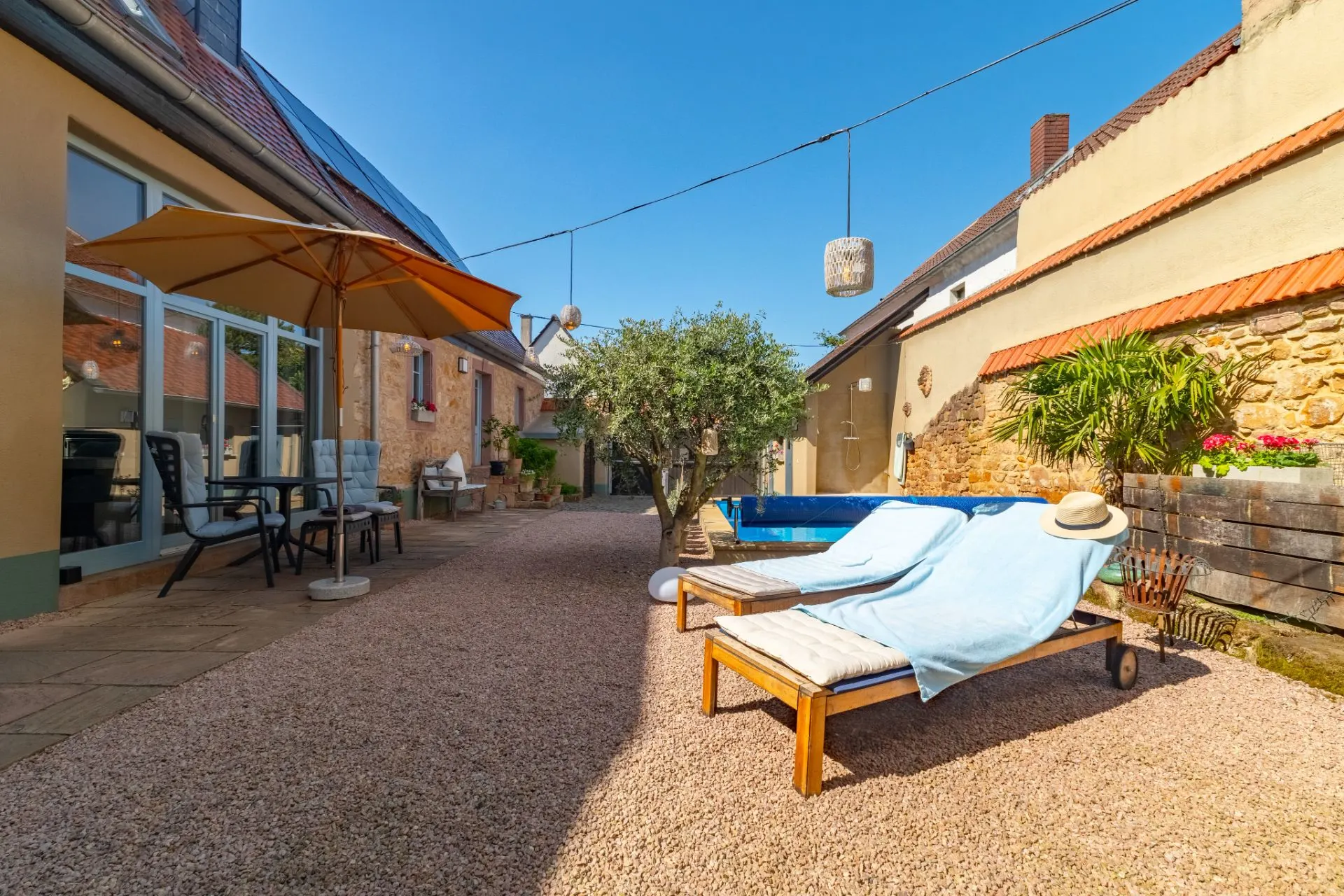 Hof mit Sonnenliegen, Sonnenschirm, kleinem Pool, Baum und Sitzbereich auf Kiesboden.