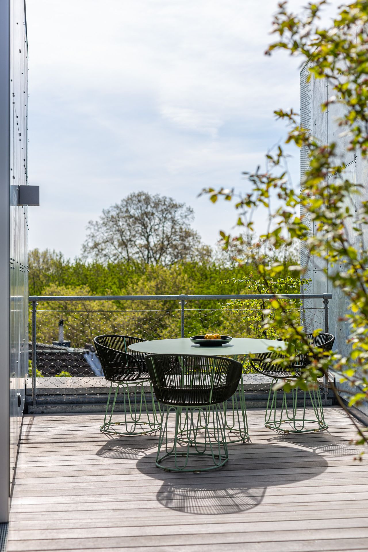 Terrasse mit rundem Tisch, vier Stühlen und einer Schale mit Obst, im Hintergrund grüne Bäume.