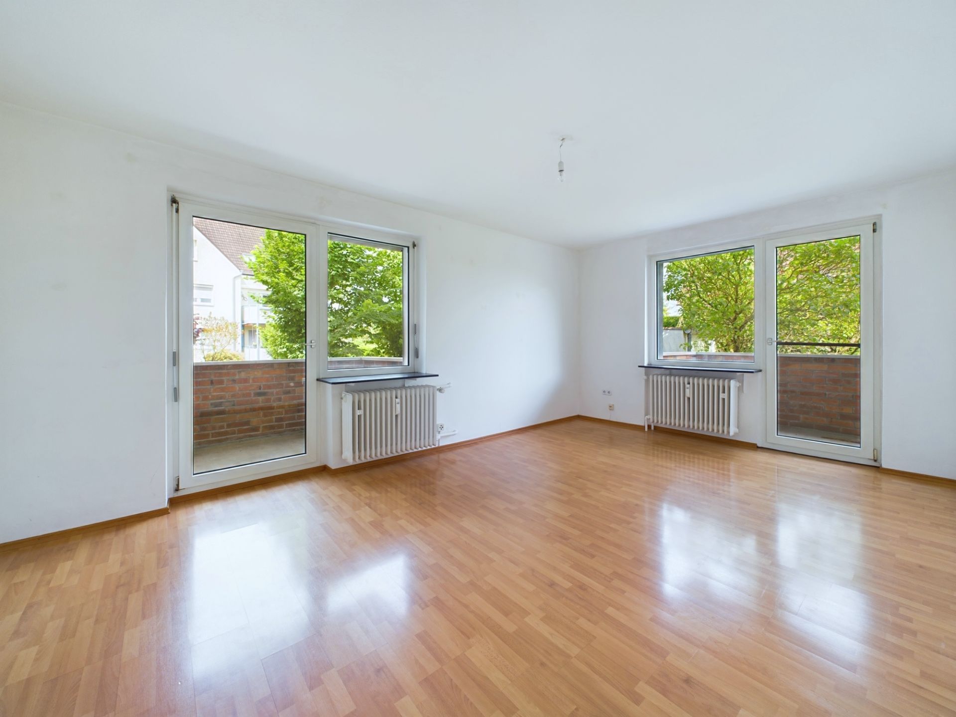 Heller leerer Raum mit Holzfußboden, zwei Fenstern und zwei Türen zum Balkon.