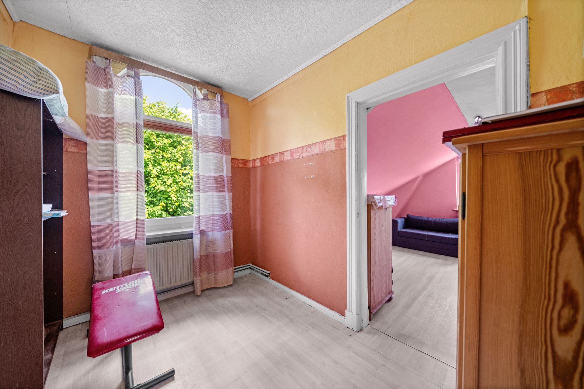Kleines Zimmer mit Fenster, gestreiften Vorhängen, Stuhl und Blick in ein weiteres Zimmer.