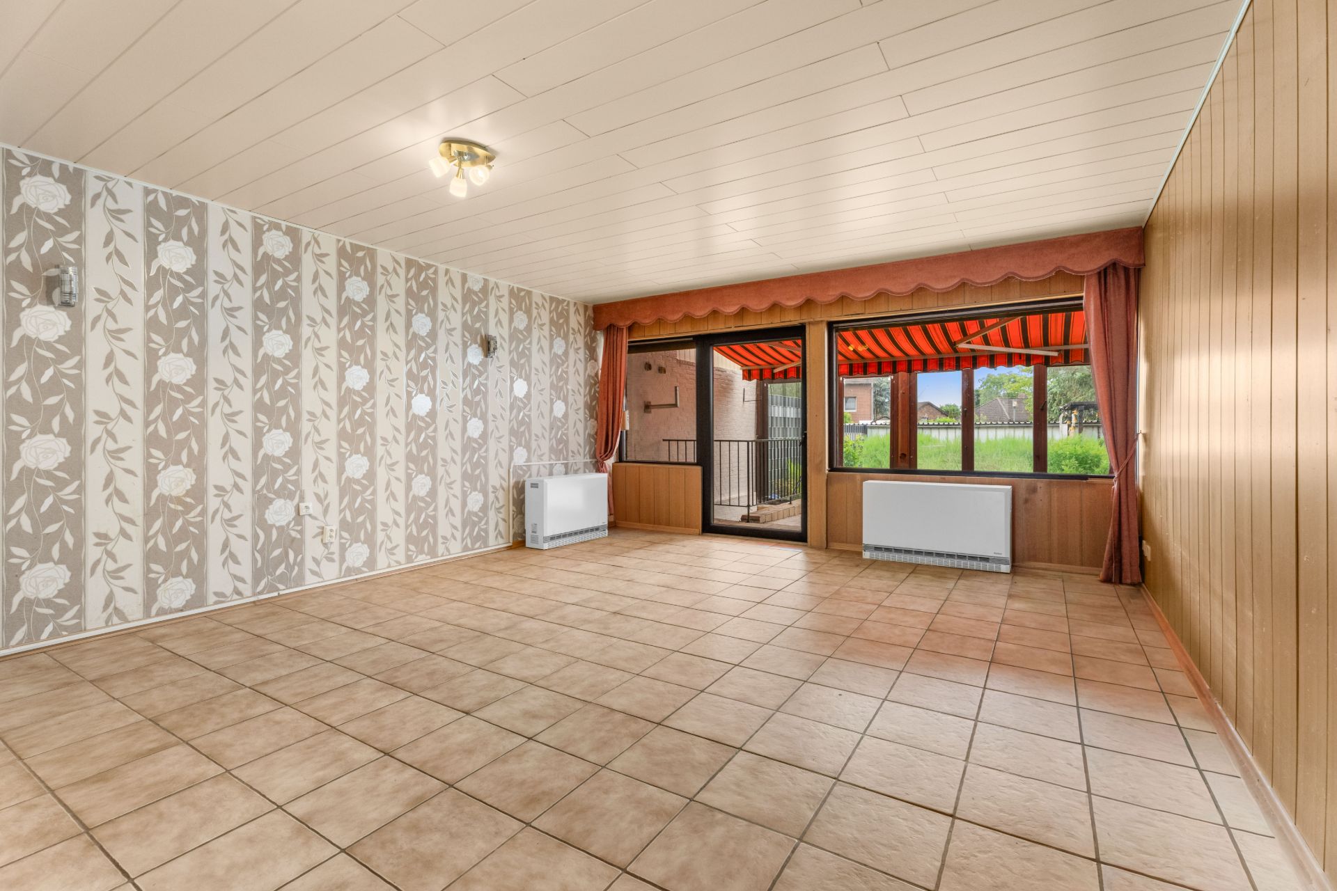Leeres Wohnzimmer mit gemusterter Tapete, Fliesenboden und großen Fenstern mit Blick auf den Garten.