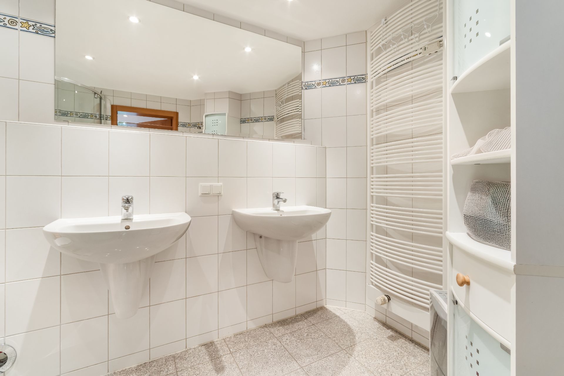 Helles Badezimmer mit zwei Waschbecken, großem Spiegel, Handtuchheizkörper und Regal.