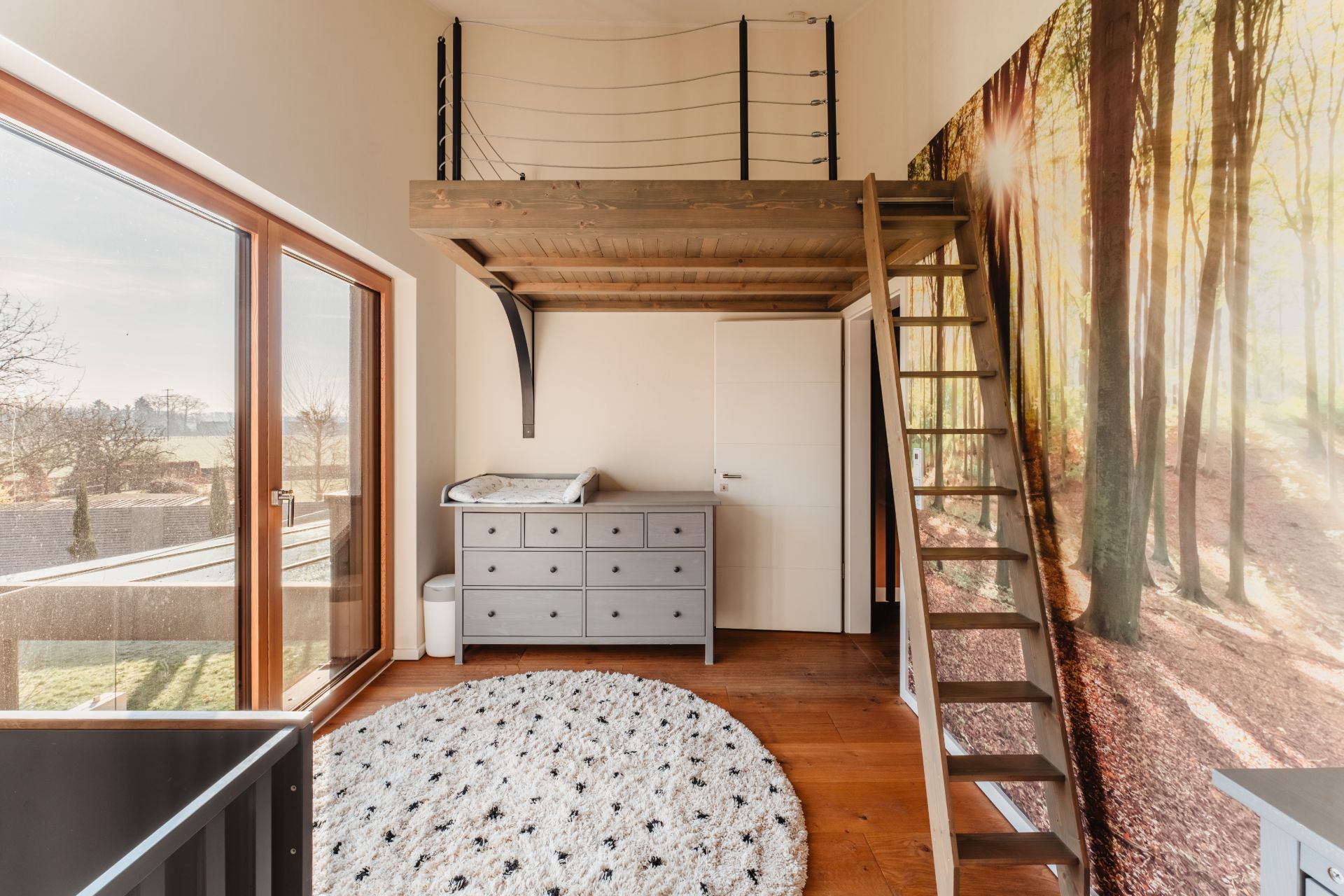 Helles Zimmer mit Hochbett, Leiter, Kommode und großem Fenster, Wand mit Waldmotiv.