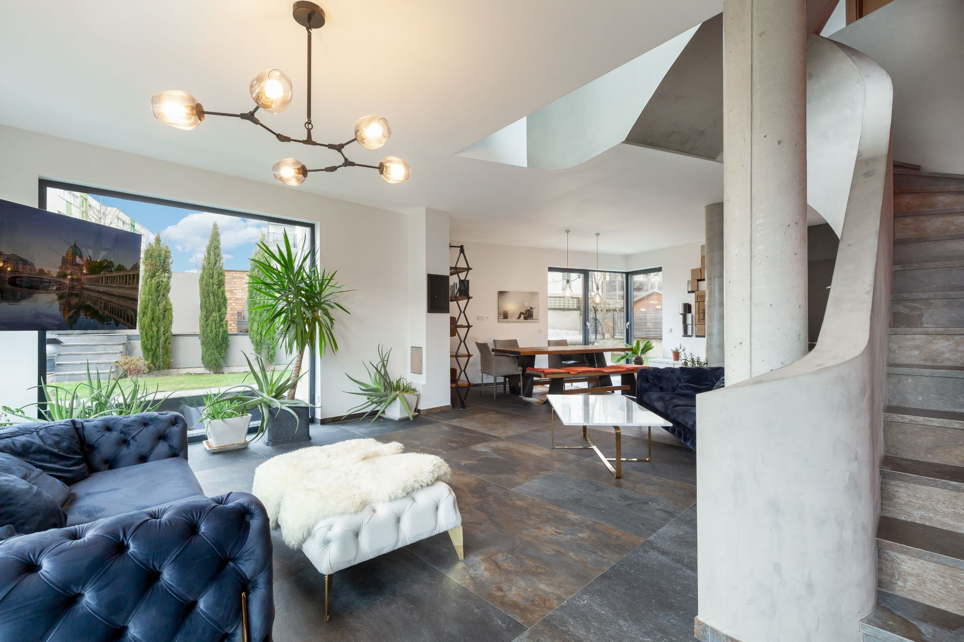 Helles Wohn- und Esszimmer mit Pflanzen, blauen Sofas, großem Fenster und moderner Treppe.