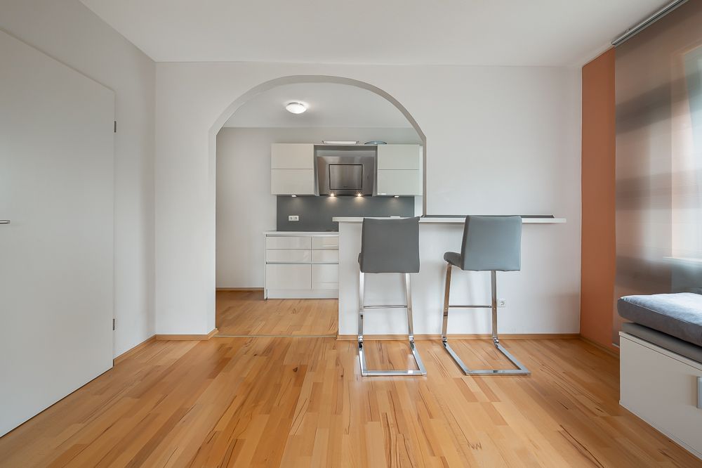 Moderne kleine Küche mit Tresen und zwei grauen Barhockern, Holzfußboden, helles Fenster.