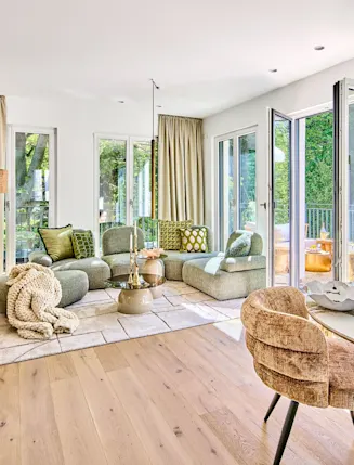 Modernes Wohnzimmer mit Sofa, Sessel, Glastisch, Essbereich und großen Fenstern zum Balkon.
