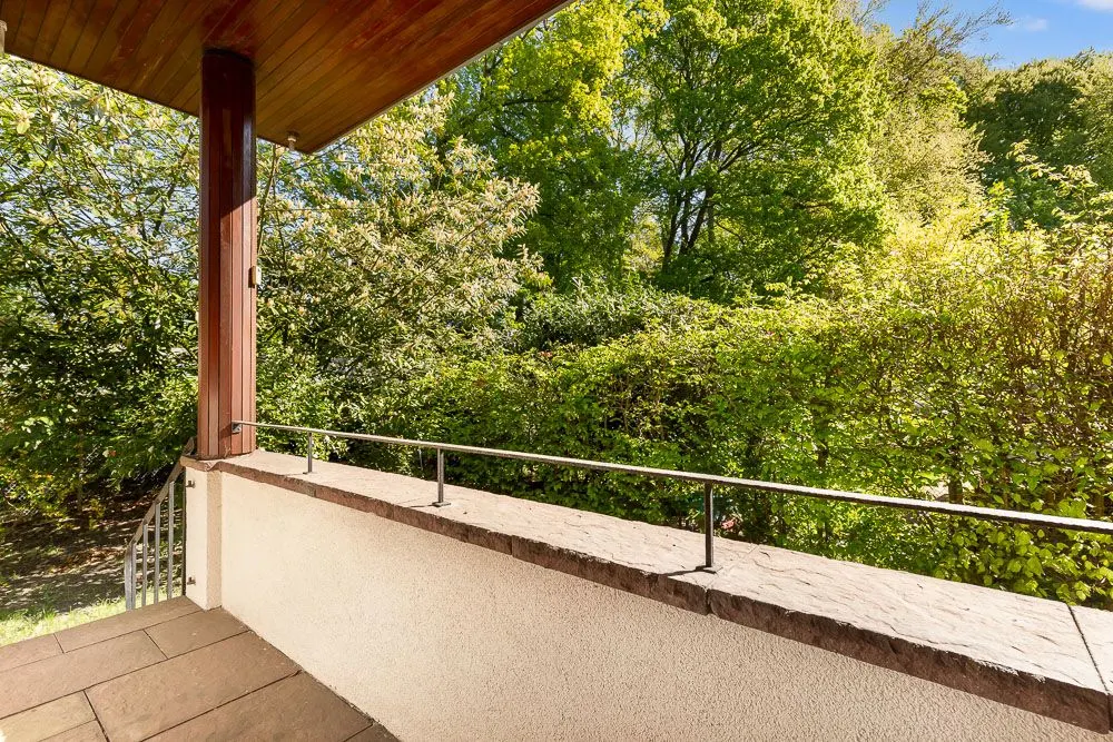 Überdachte Terrasse mit Geländer und Blick auf dichte grüne Bäume und Sträucher.