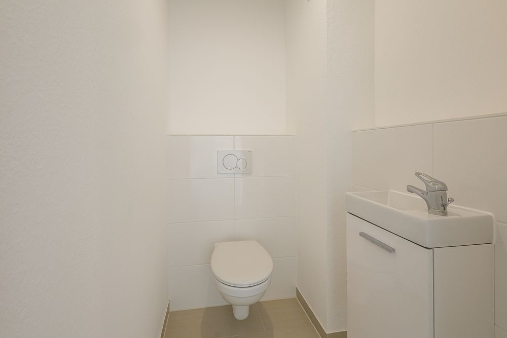 Kleines, weiß gefliestes WC mit Wandtoilette und kleinem Waschbecken.
